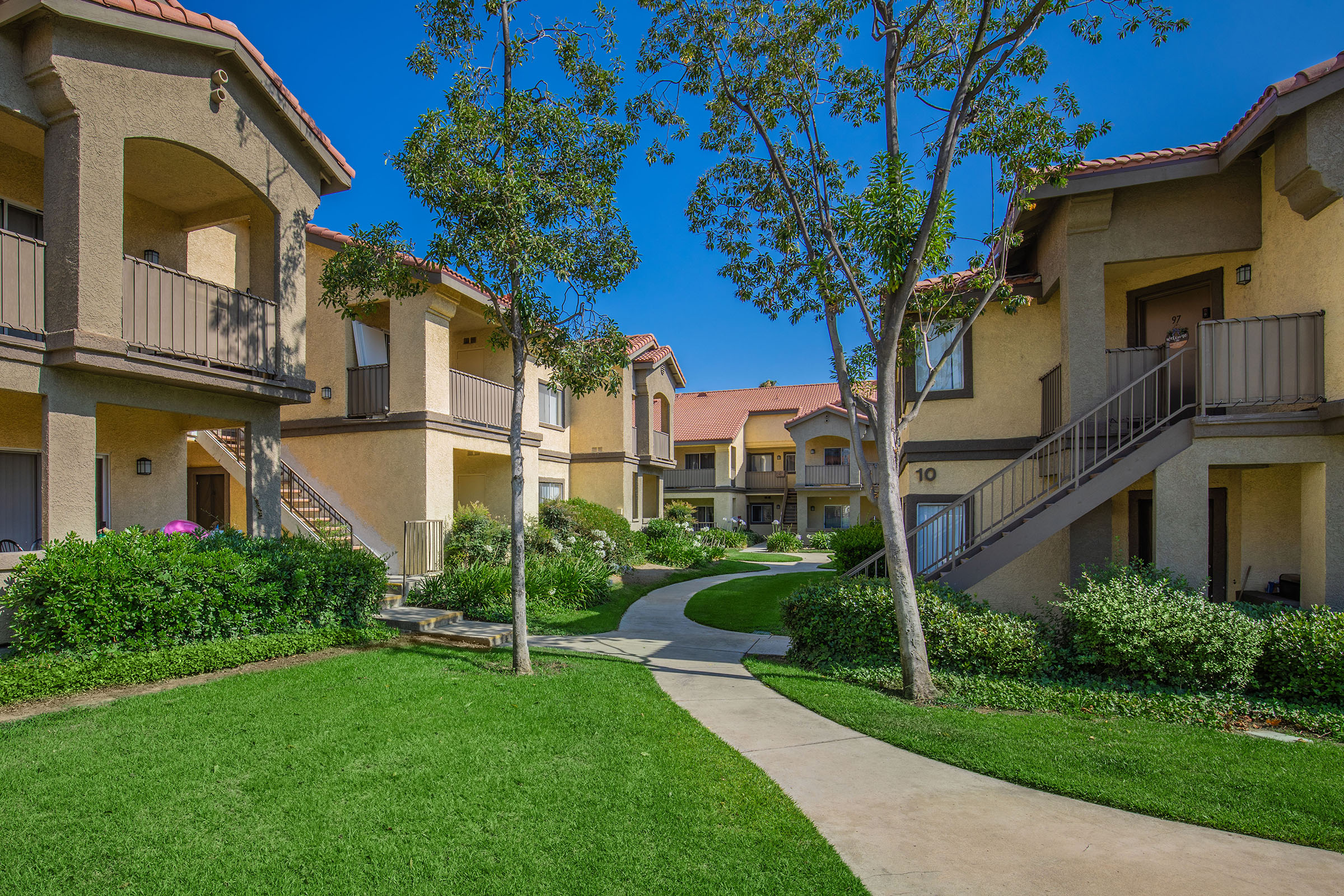 A landscaped courtyard with sidewalks and green grass, surrounded by residential apartment buildings. Small trees and shrubbery enhance the outdoor space, with a clear blue sky overhead. The scene conveys a serene and inviting atmosphere in a neighborhood setting.