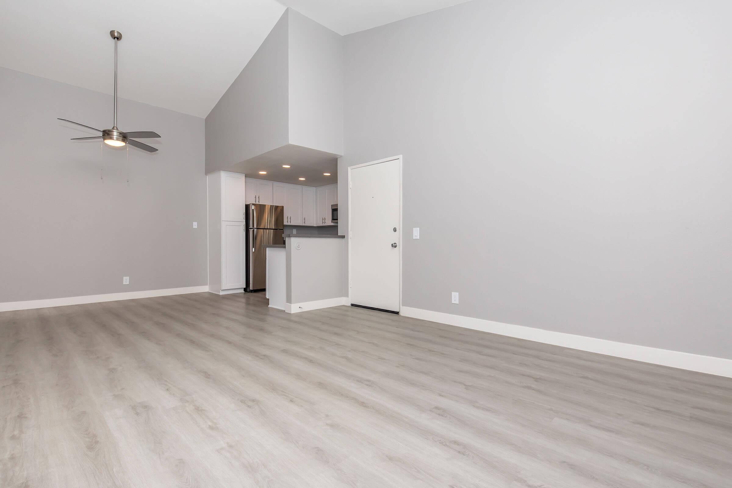 A spacious, modern living area featuring light gray walls, laminate flooring, and high ceilings. A ceiling fan is mounted above, and to the left, there is an open kitchen area with stainless steel appliances visible. The entrance door is on the right side, leading outside.