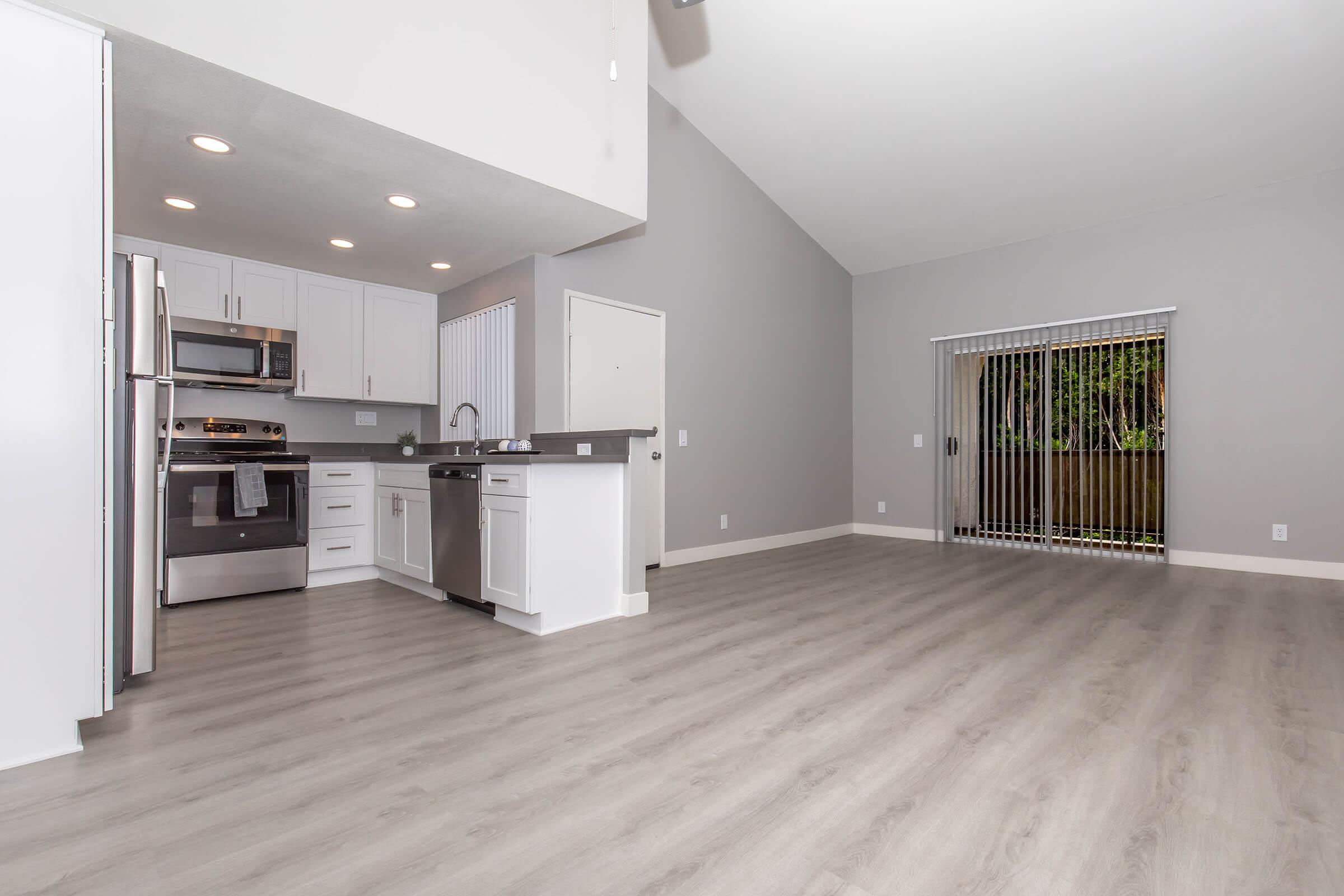 A modern kitchen and living area featuring stainless steel appliances, white cabinetry, and gray walls. The open layout includes a spacious floor with light wood-like flooring. Large windows with blinds provide natural light, and there is a door leading to an outdoor space.