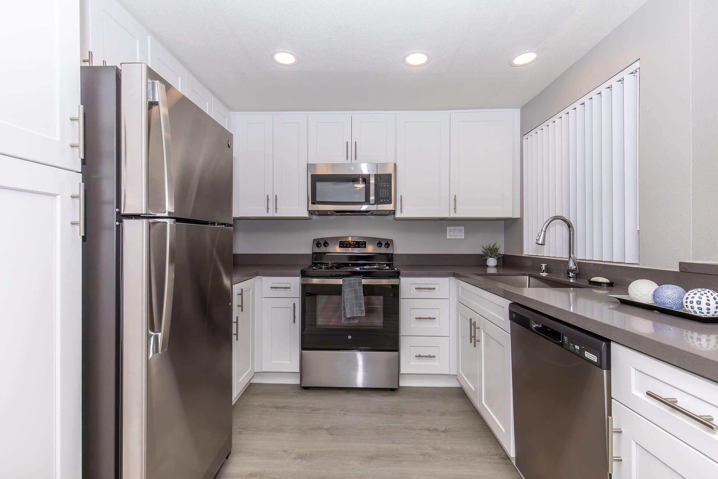 Modern kitchen featuring stainless steel appliances, including a refrigerator, microwave, and oven. The cabinetry is white with a sleek countertop. There are tight blinds on the window and decorative items on the countertop. The design is clean and contemporary, with ample lighting from recessed fixtures.