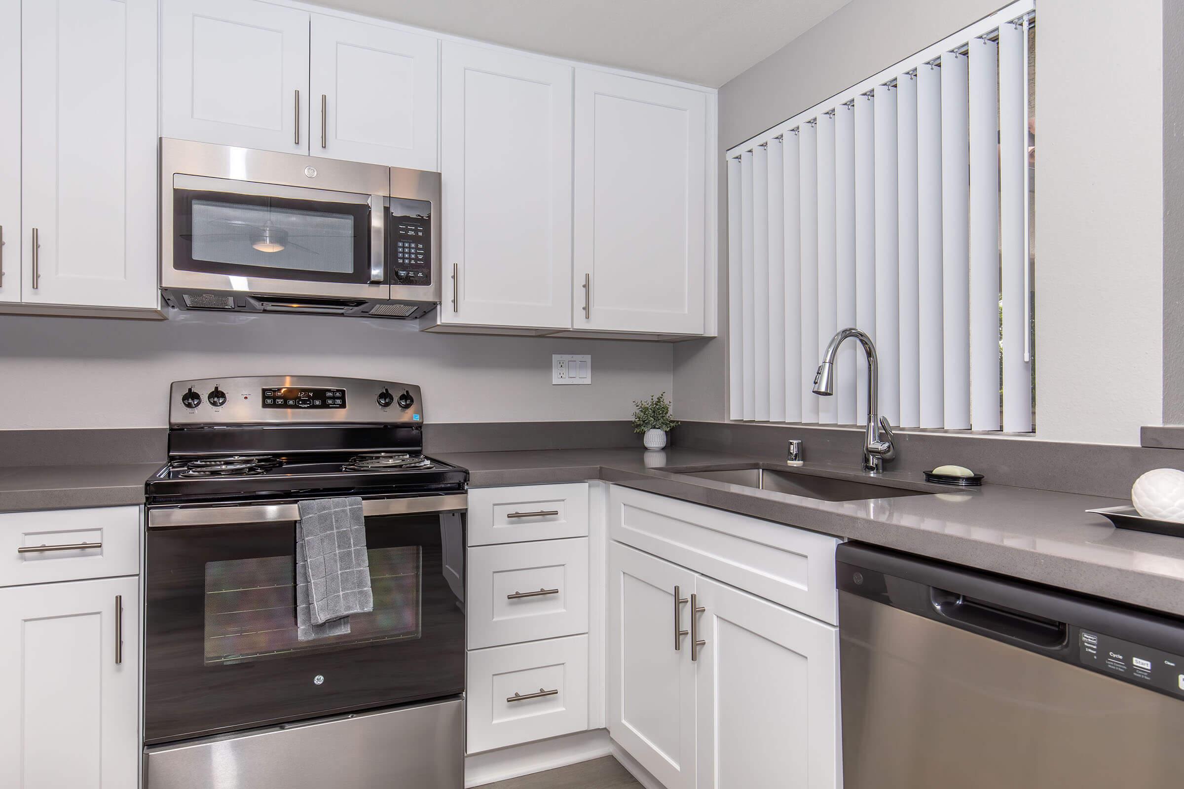 Modern kitchen featuring white cabinetry, stainless steel appliances including a microwave, oven, and dishwasher, a gray countertop, and a sink with a sleek faucet. The space is well-lit, with a window covered by horizontal blinds, and a small plant adds a touch of greenery.