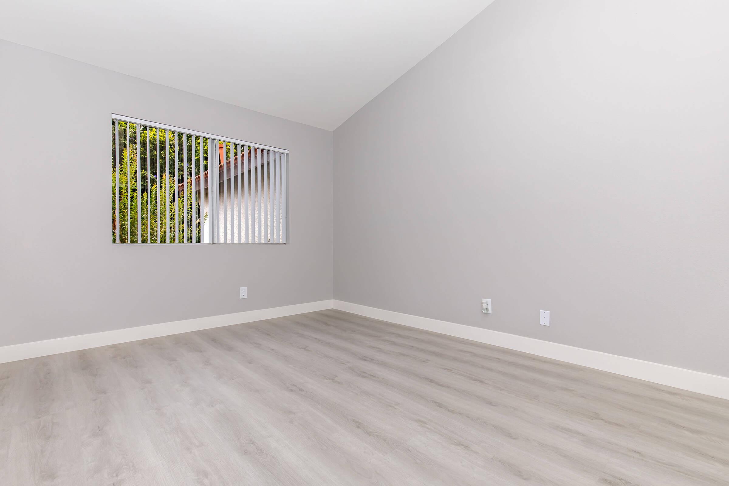 A spacious, empty room with light gray walls and a high ceiling. Vinyl flooring in a light tone covers the floor. A window with vertical blinds is situated on the left side, allowing natural light to brighten the space. The overall look is clean and minimalistic, ideal for various uses.