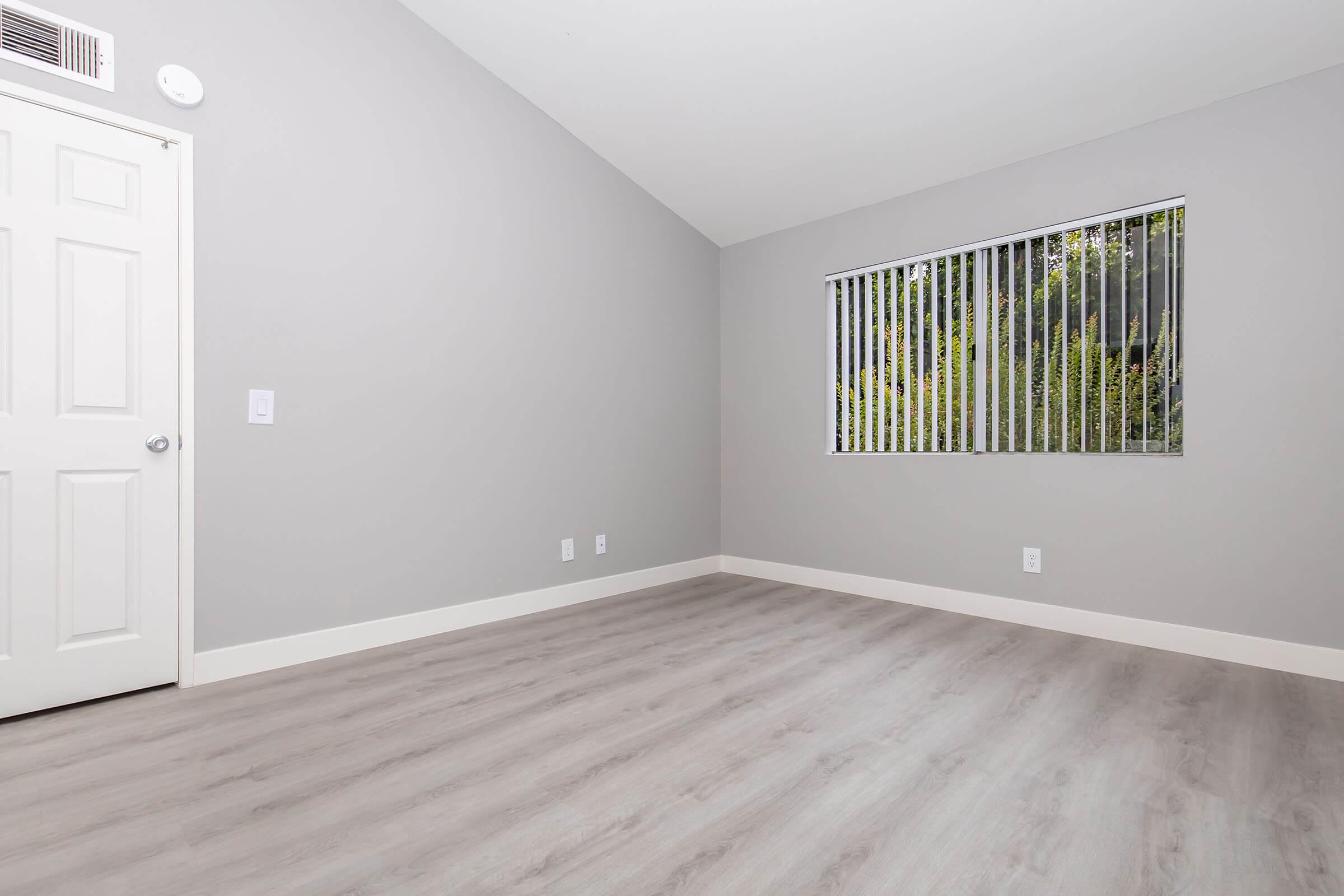 A sparsely furnished room featuring light gray walls and a light wood floor. The room has a door on the left, a window with vertical blinds showing greenery outside, and no visible furniture or decorations, creating a clean and minimalist appearance.
