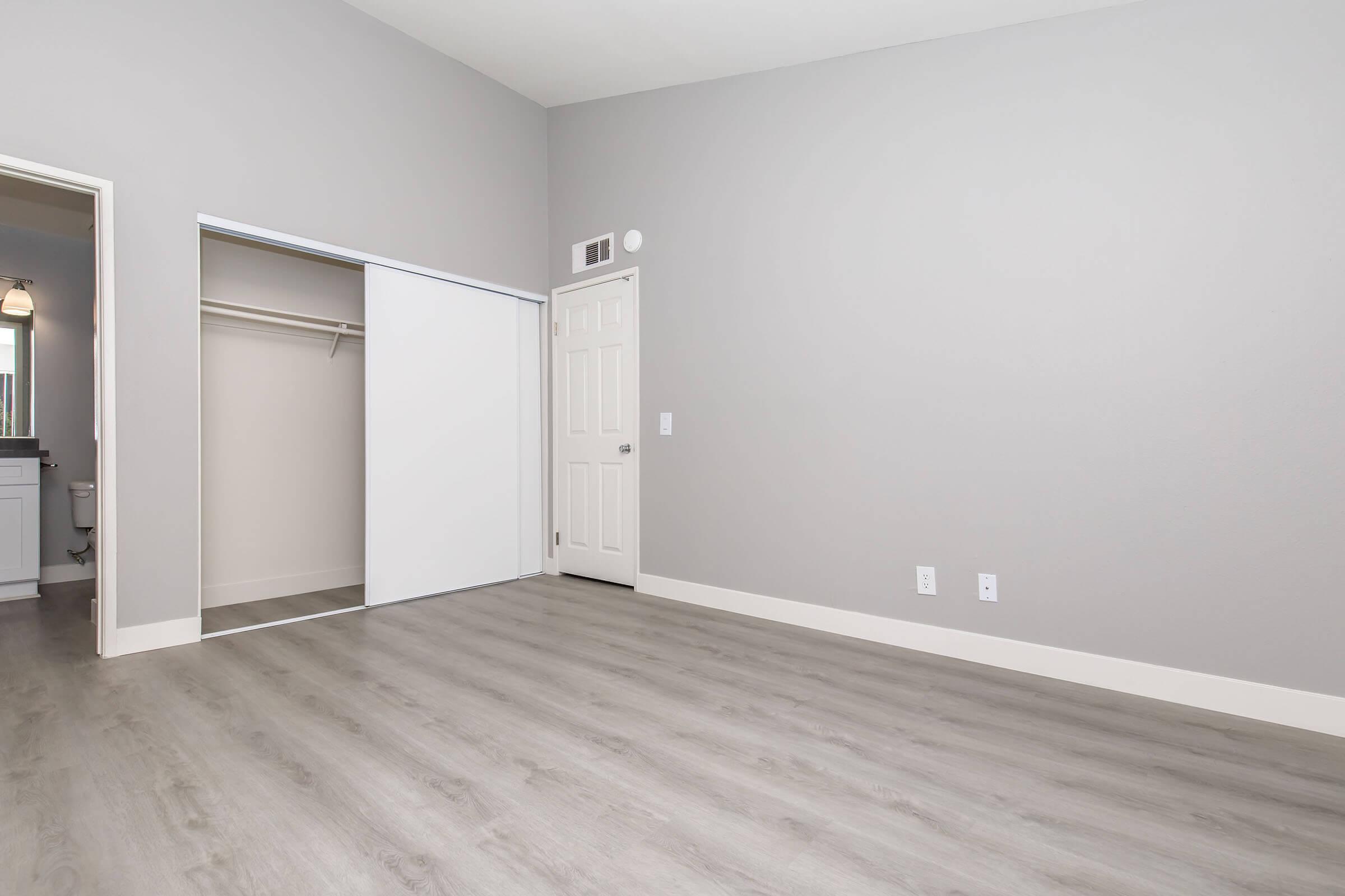 A spacious, empty room with gray walls and a light wood floor. On one side, there is a sliding closet door, and a door leading to another area is visible. Natural light streams in, creating a bright and airy atmosphere. The room has a clean and modern aesthetic, perfect for various uses.