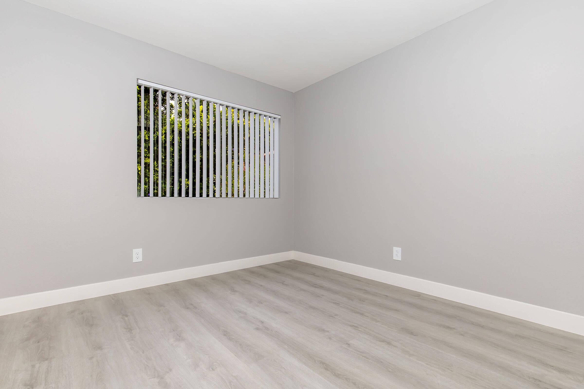 A minimalist empty room with light gray walls, a sliding window covered by vertical blinds, and wooden flooring. The space is bright and airy, with no furniture present, creating a blank canvas for decoration or design.
