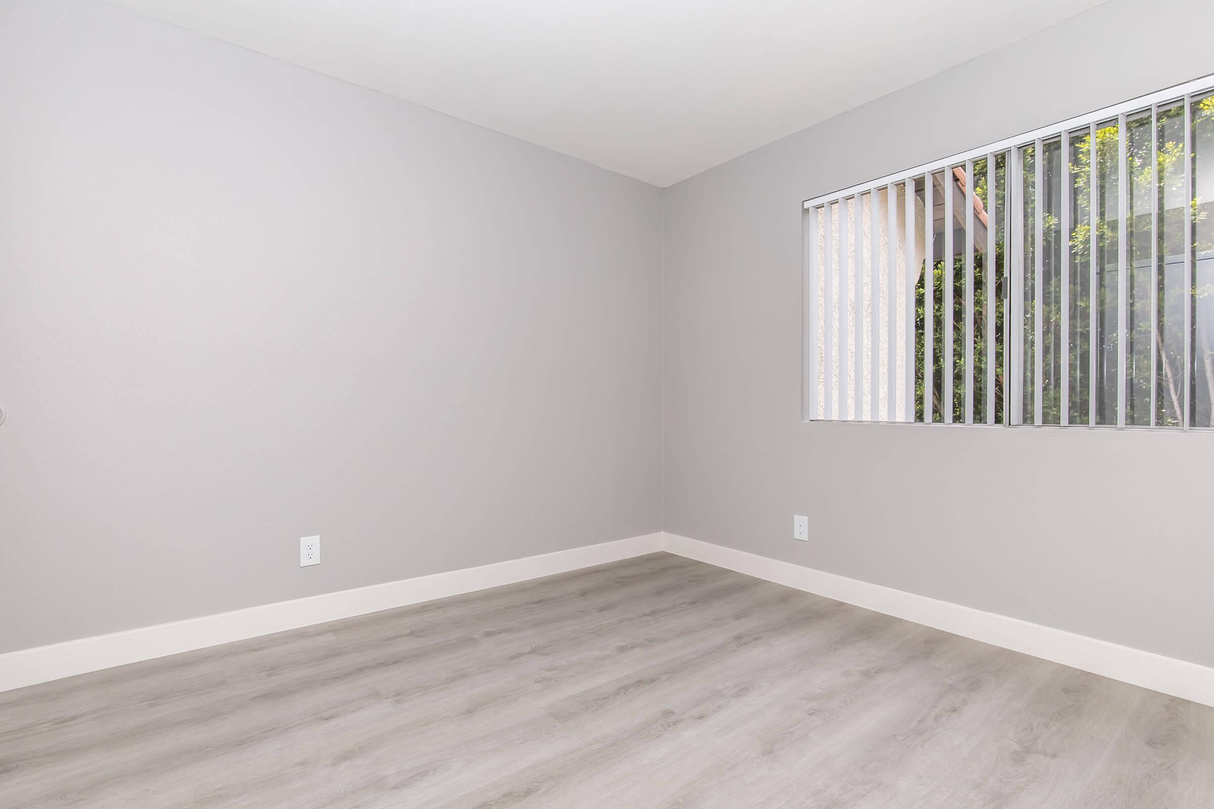 Empty room with light gray walls and laminate flooring. A window with vertical blinds lets in natural light, and there are no furnishings or decorations present. The overall atmosphere is clean and minimalistic.