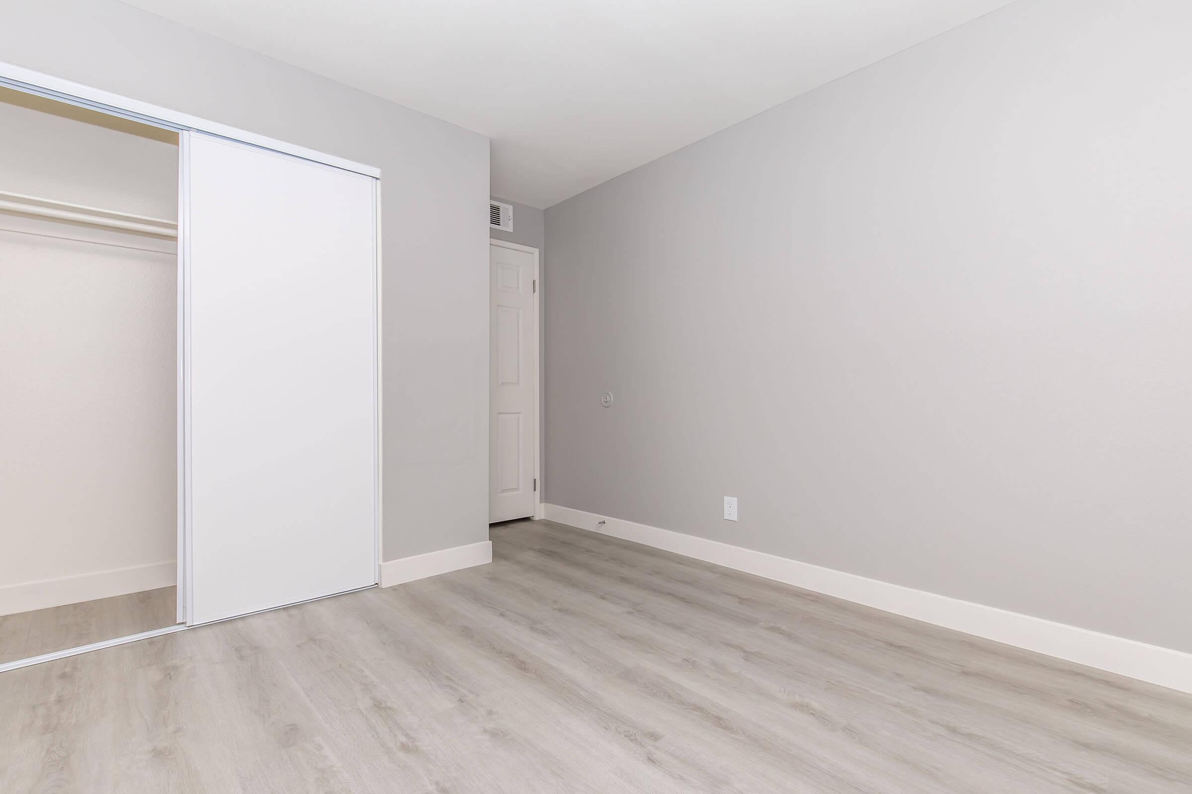 A light gray room with a smooth wooden floor. There is a white sliding closet door on the left and a door leading to another room on the right. The walls are bare, creating a minimalist and spacious feel. Natural light brightens the space, enhancing the overall airy atmosphere.