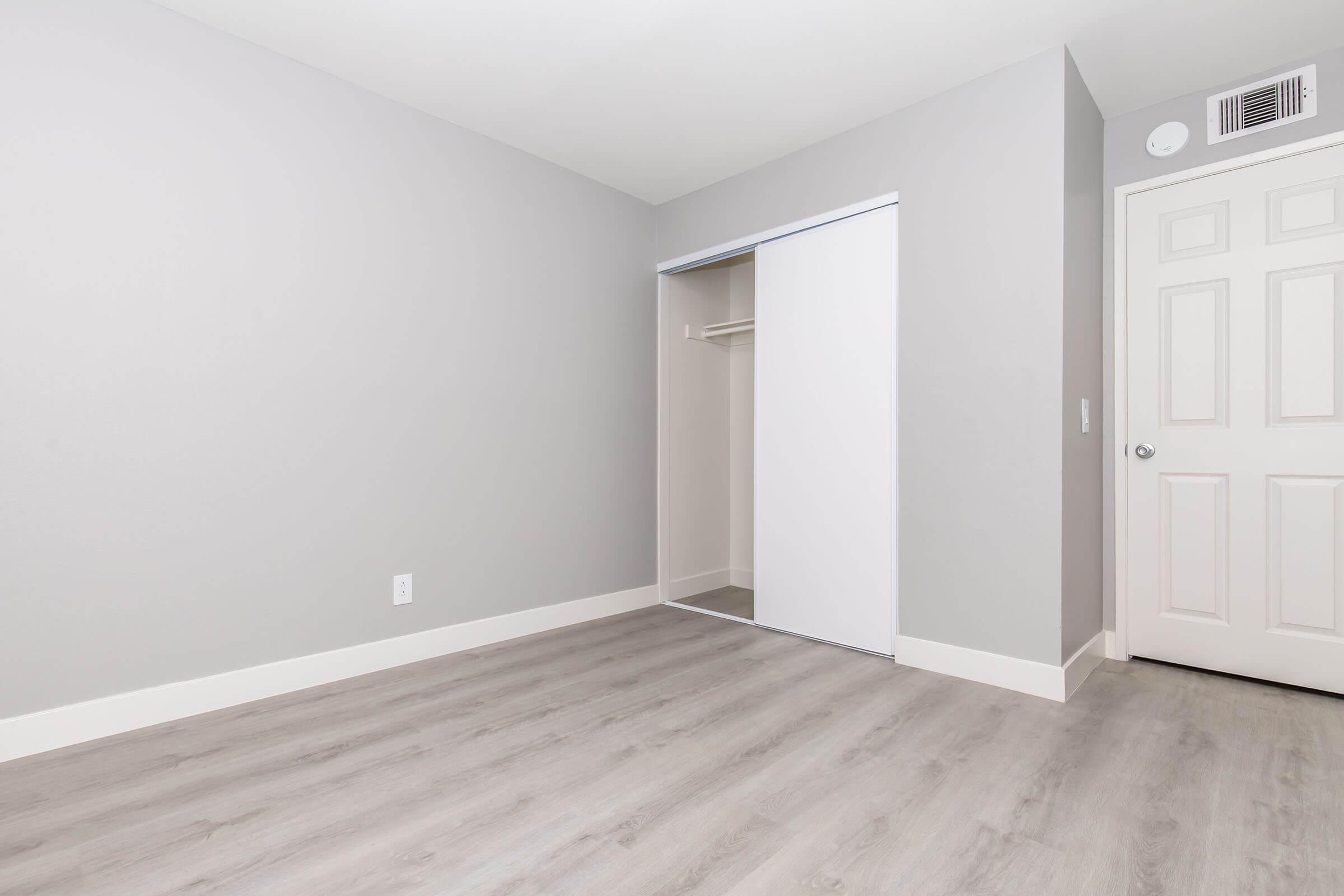Empty room with light gray walls and a light gray laminate floor. A sliding closet door is visible on the left, and a white door is on the right. The space is well-lit and minimalistic, suitable for various uses such as a bedroom or office. No furniture is present.