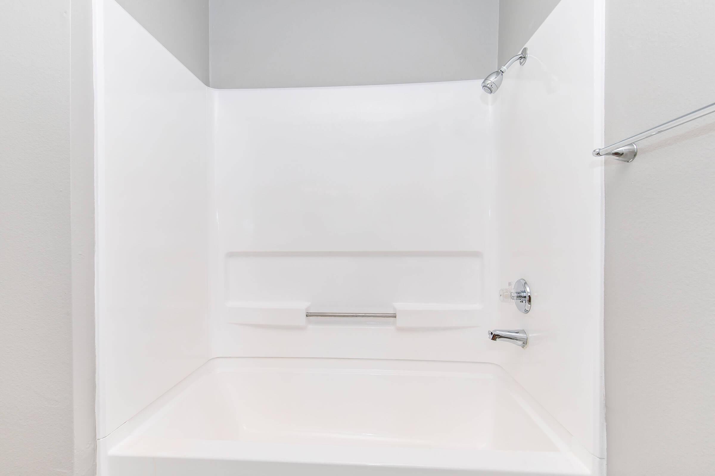 A clean, modern bathtub with a curved, white shower surround. The space is well-lit, with a chrome showerhead and a single faucet on the wall. A grab bar is mounted for safety. The walls are painted a light color, complementing the simplicity of the design.