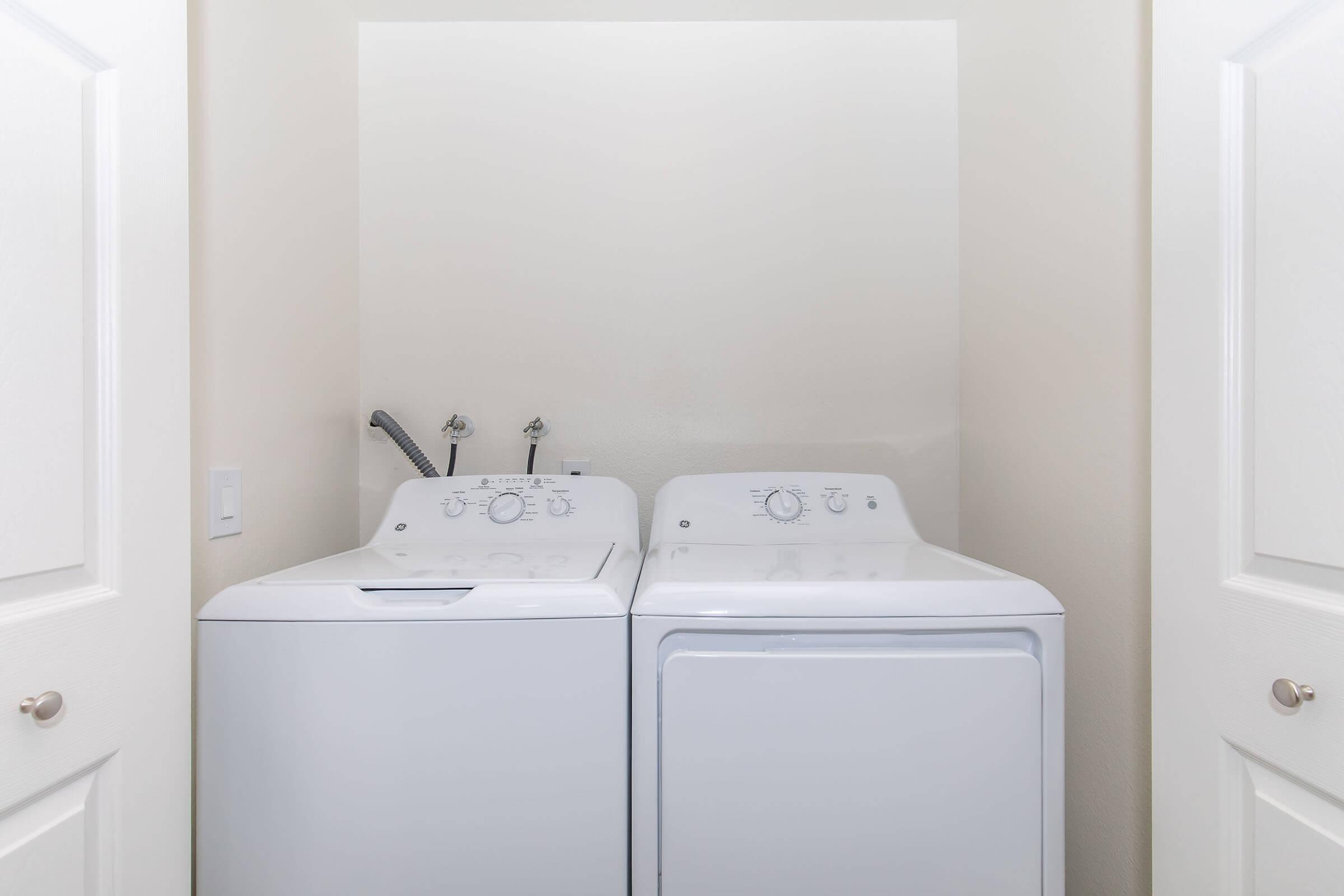 A white washing machine and dryer are placed side by side in a small laundry area. The room has light-colored walls, and there are two closed white doors visible, framing the appliances. The appliances feature control dials and a hose attached to the washing machine.