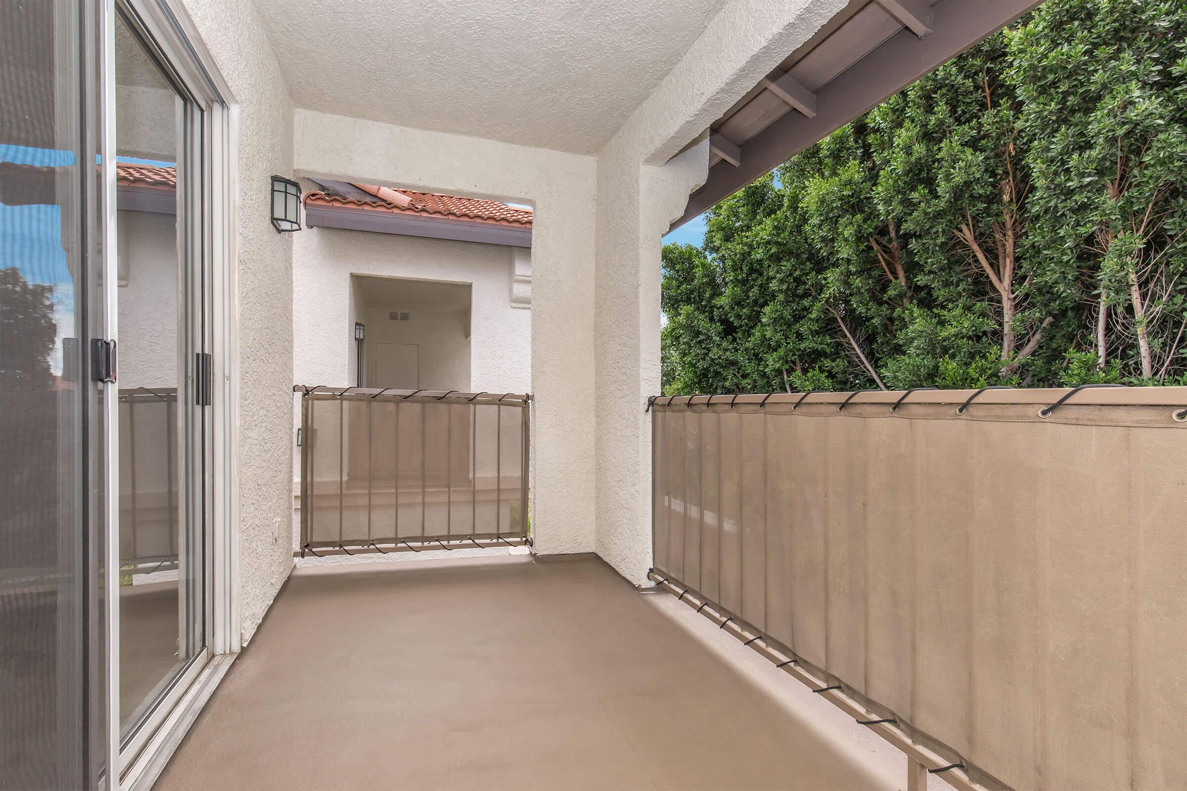 A spacious balcony with a beige railing overlooking greenery, featuring a textured floor and walls. Large sliding glass doors provide access, and there is a light fixture on the wall. The area is bright and inviting, suggesting a peaceful outdoor space.
