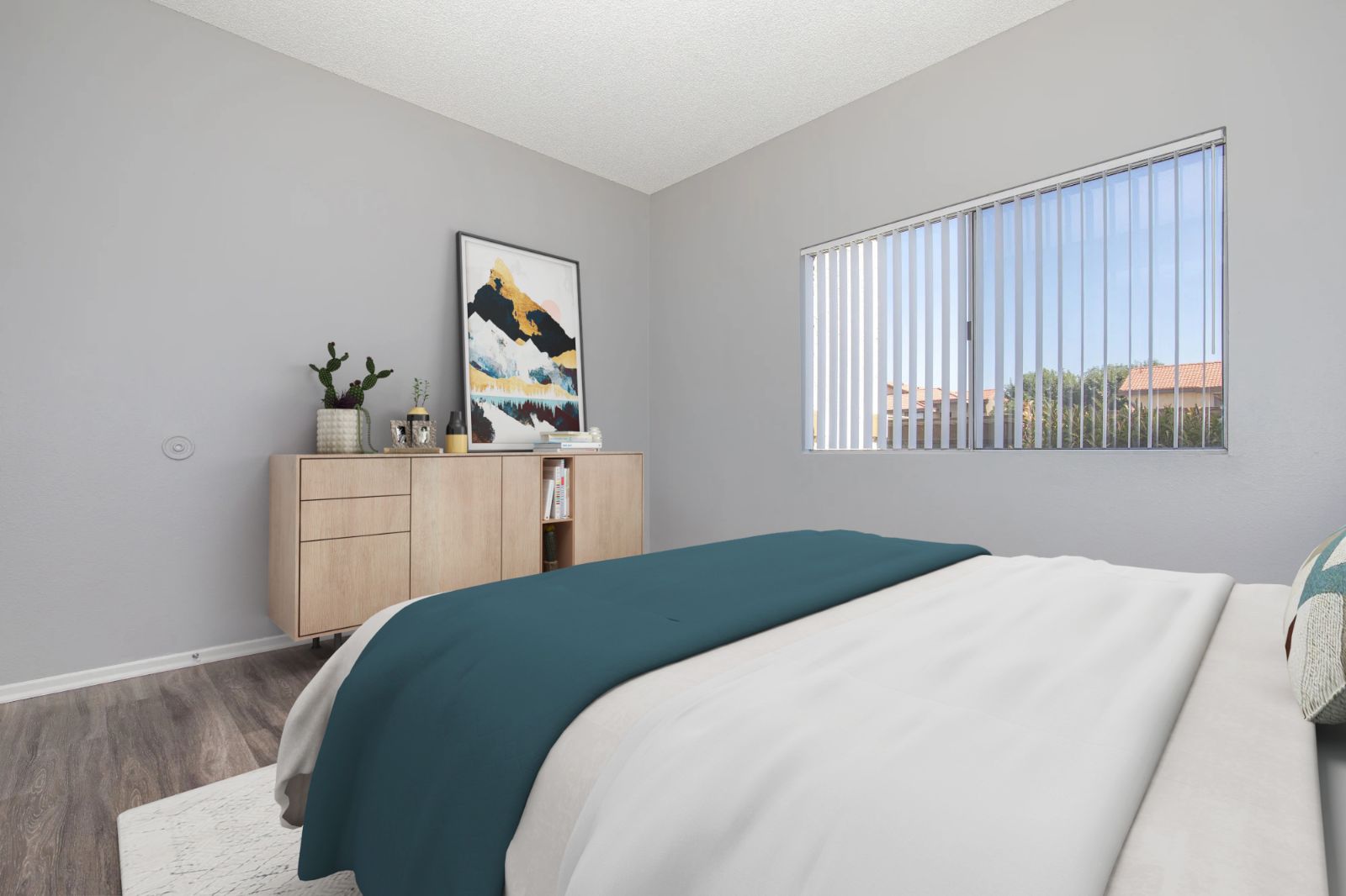 A neatly arranged bedroom featuring a large window with vertical blinds, allowing natural light to enter. The bed is covered with a teal and white comforter. A wooden sideboard with decorative items stands against the wall, and a piece of abstract art is displayed above it. A small plant adds a touch of greenery to the room.