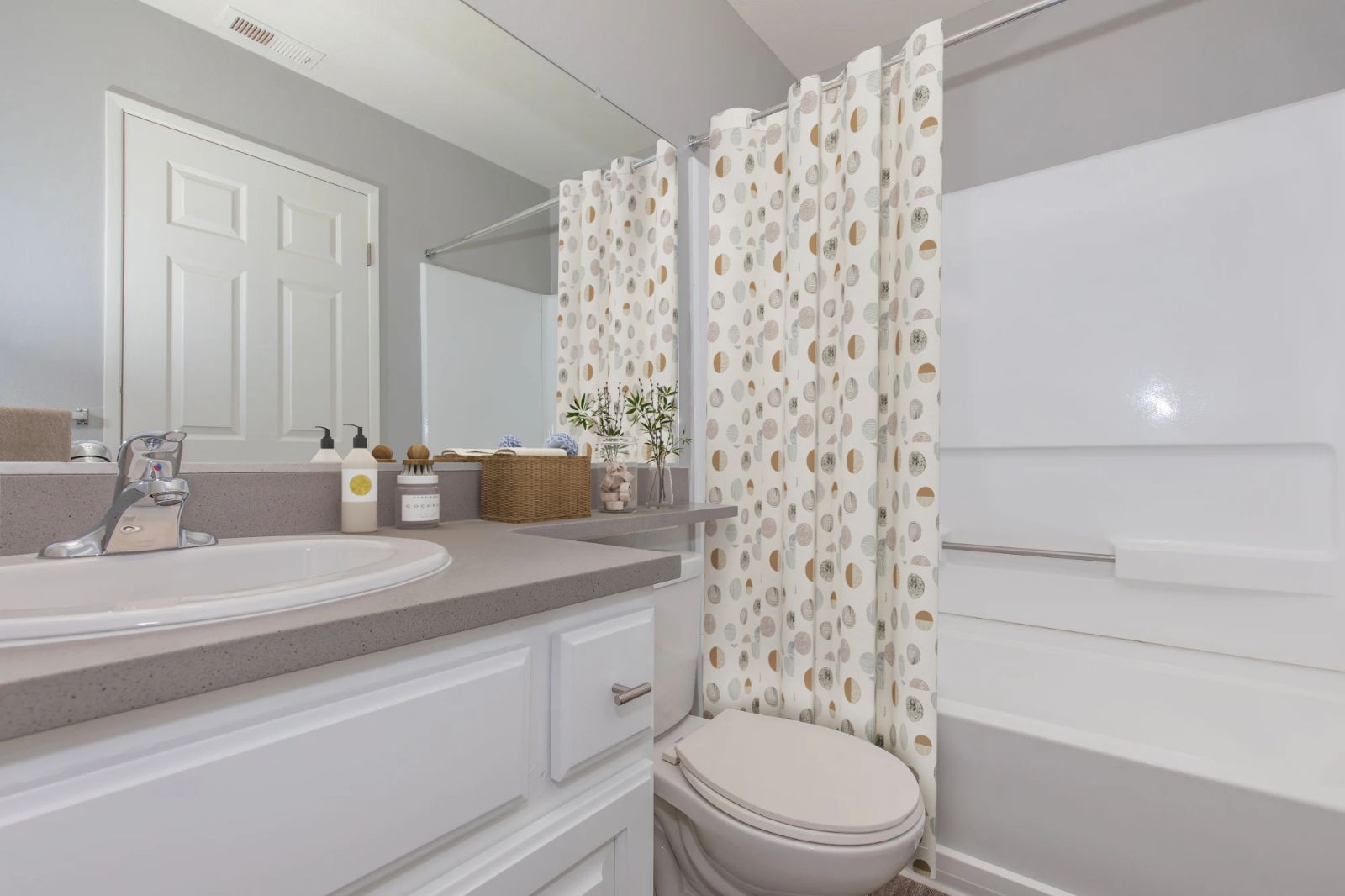 A clean, modern bathroom featuring a countertop with sink, a mirror, and a shower area with a patterned curtain. The space includes toiletries on the counter and a small potted plant, with light gray walls and a white bathtub. The door is closed in the background.