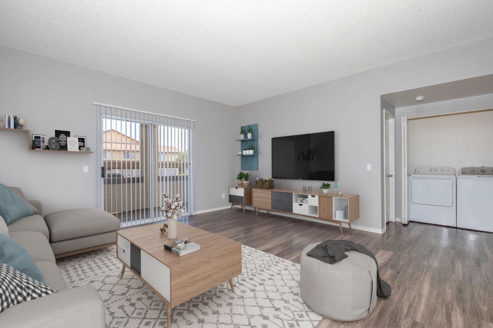 A modern living room featuring a light gray wall and flooring. There is a beige sofa, a wooden coffee table, and a large TV mounted on the wall. A sliding door leads to a balcony, and shelves with decor are visible. In the background, a laundry area can be seen through an open door.