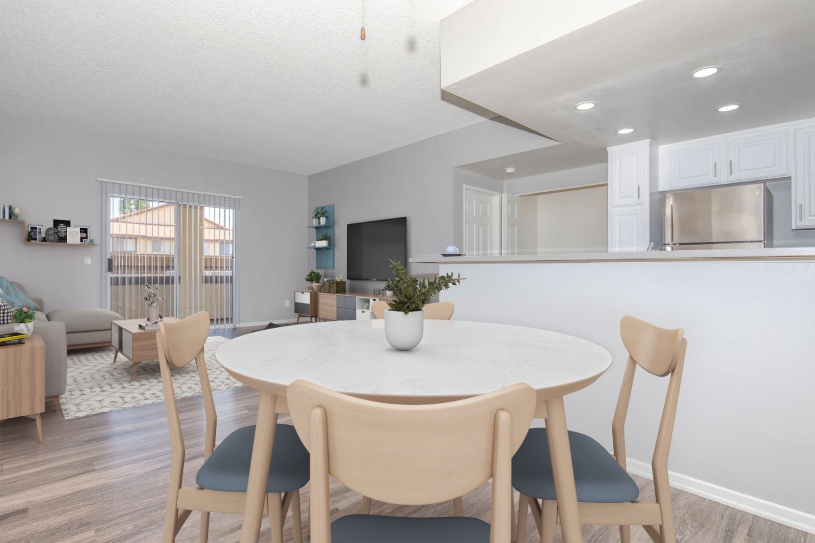 A modern dining area featuring a round marble table surrounded by wooden chairs, with a view of a bright living room and kitchen in the background. The space has light gray walls and wooden flooring, with a sliding glass door leading to an outdoor area. Decorative elements and a TV are visible in the living space.