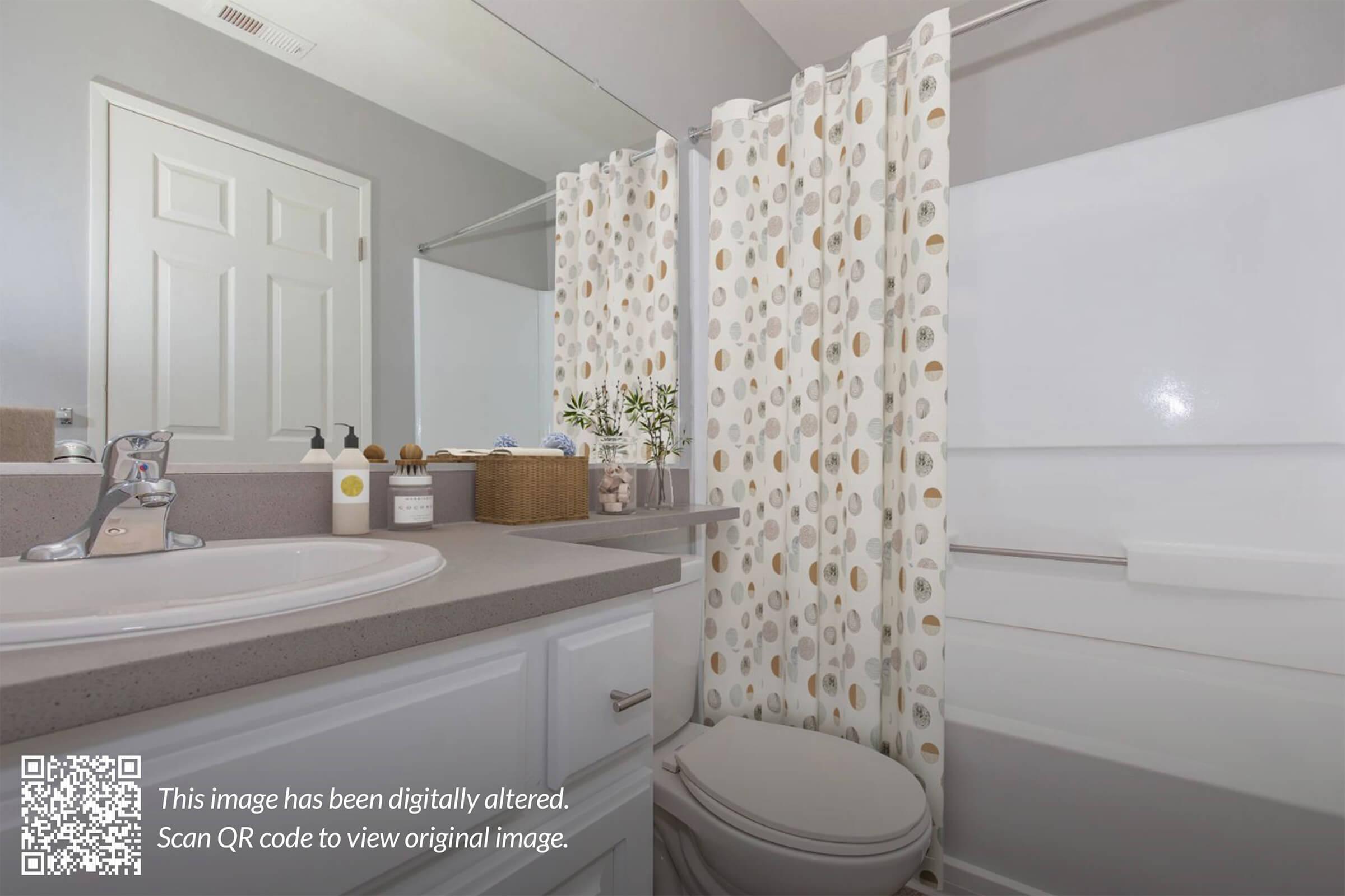 A clean, modern bathroom featuring a white toilet, sink with a silver faucet, and a gray countertop. The shower area has a clear glass door and a shower curtain with a circular pattern. A basket and decorative items are on the countertop. Bright lighting creates a fresh ambiance.