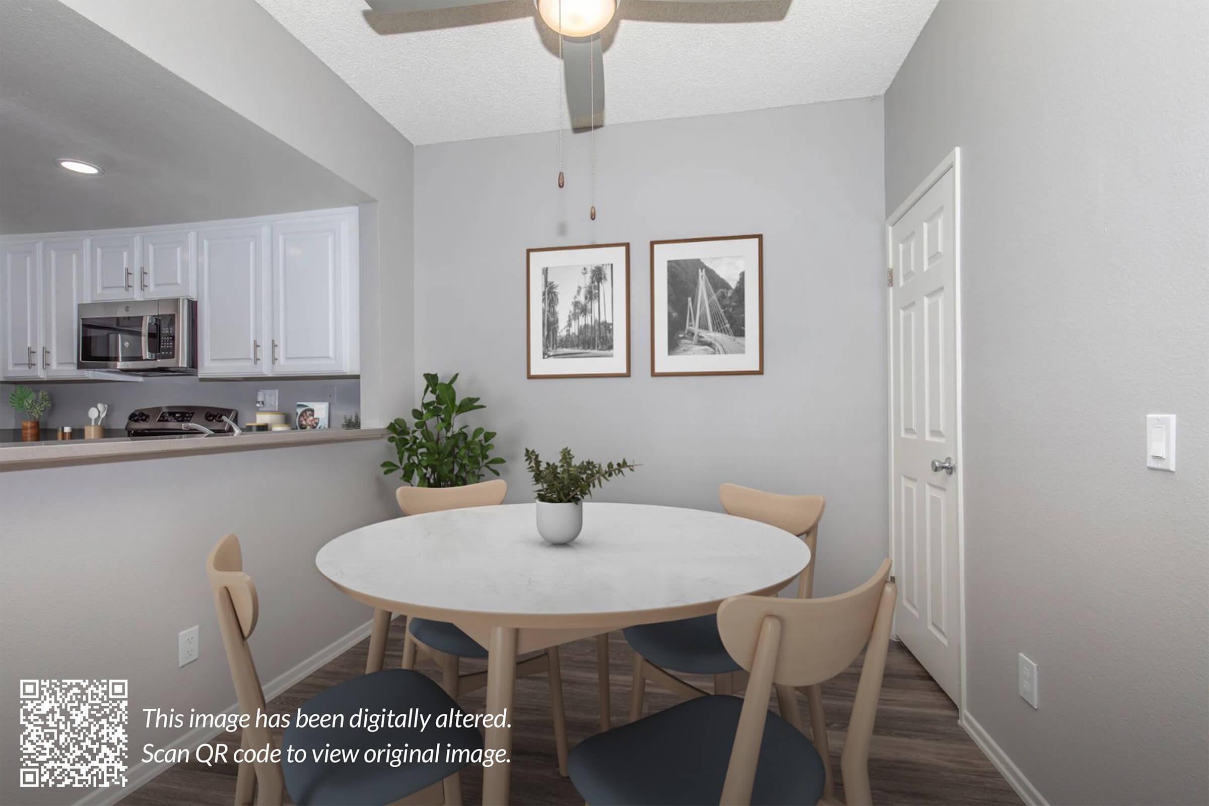 A modern dining area featuring a round marble table surrounded by wooden chairs. In the background, a kitchen with white cabinetry and appliances is visible. Two framed pictures hang on the wall, and a small plant sits in the center of the table. Soft lighting from a ceiling fan illuminates the space.