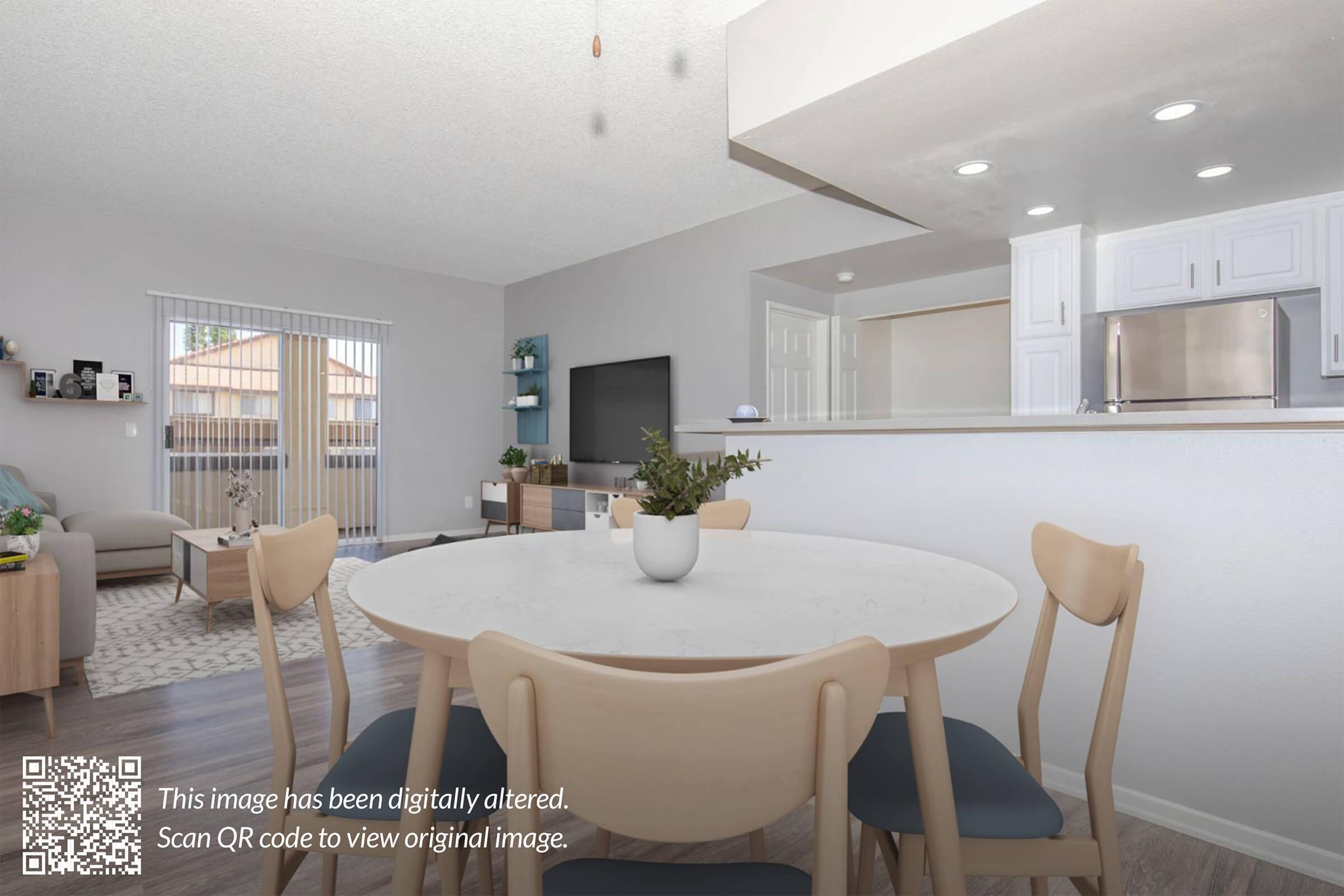 A modern dining area with a round white table surrounded by wooden chairs. In the background, a light-filled living room features a blue sofa, a TV on a stand, and large windows. The kitchen is visible with white cabinetry and stainless steel appliances, creating a bright and inviting space.