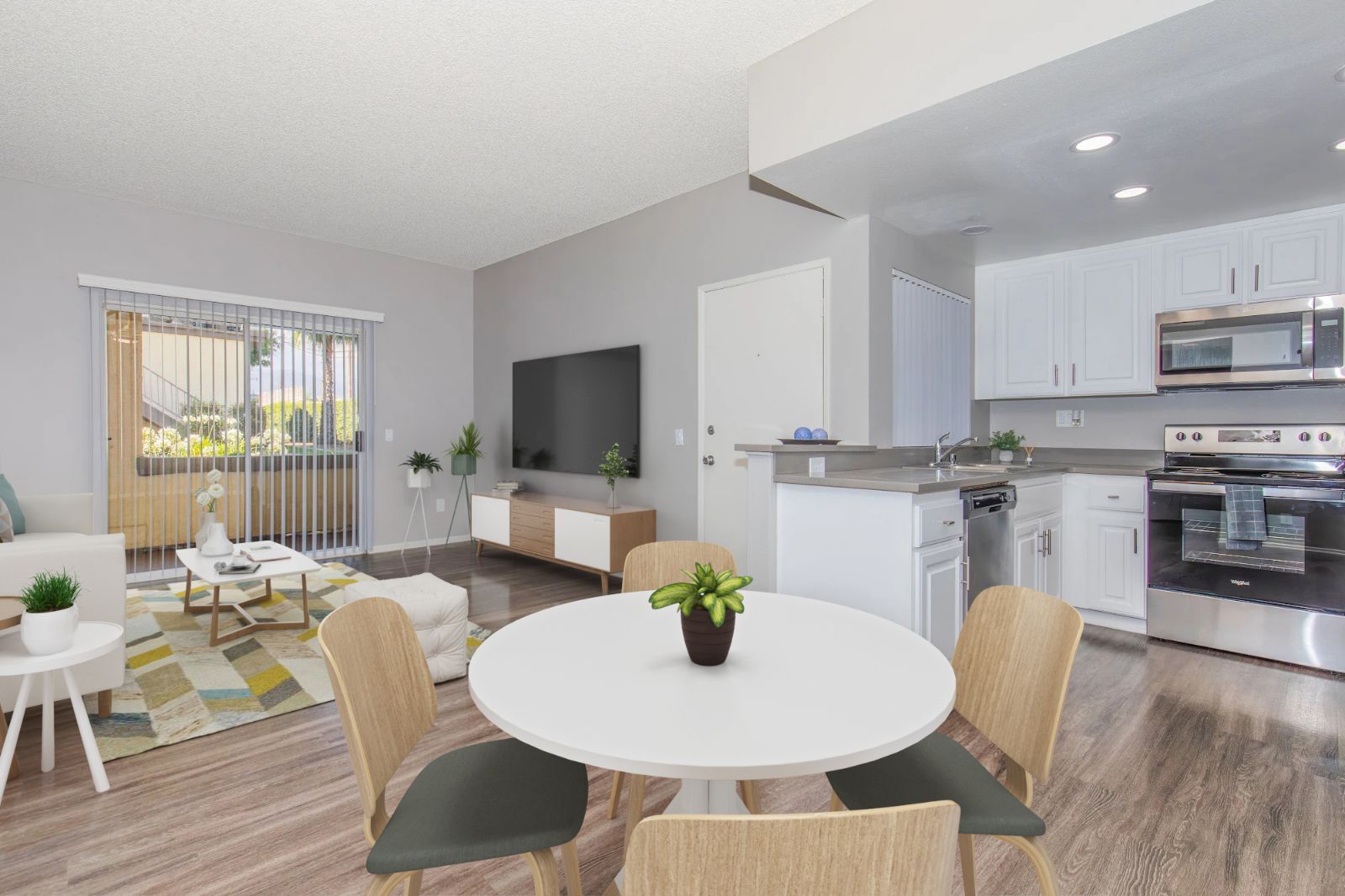 A modern open-concept living space featuring a circular dining table with four chairs, a stylish living area with a sofa and coffee table, and a kitchen with white cabinets and stainless steel appliances. Natural light streams in through sliding glass doors, enhancing the bright and inviting atmosphere.