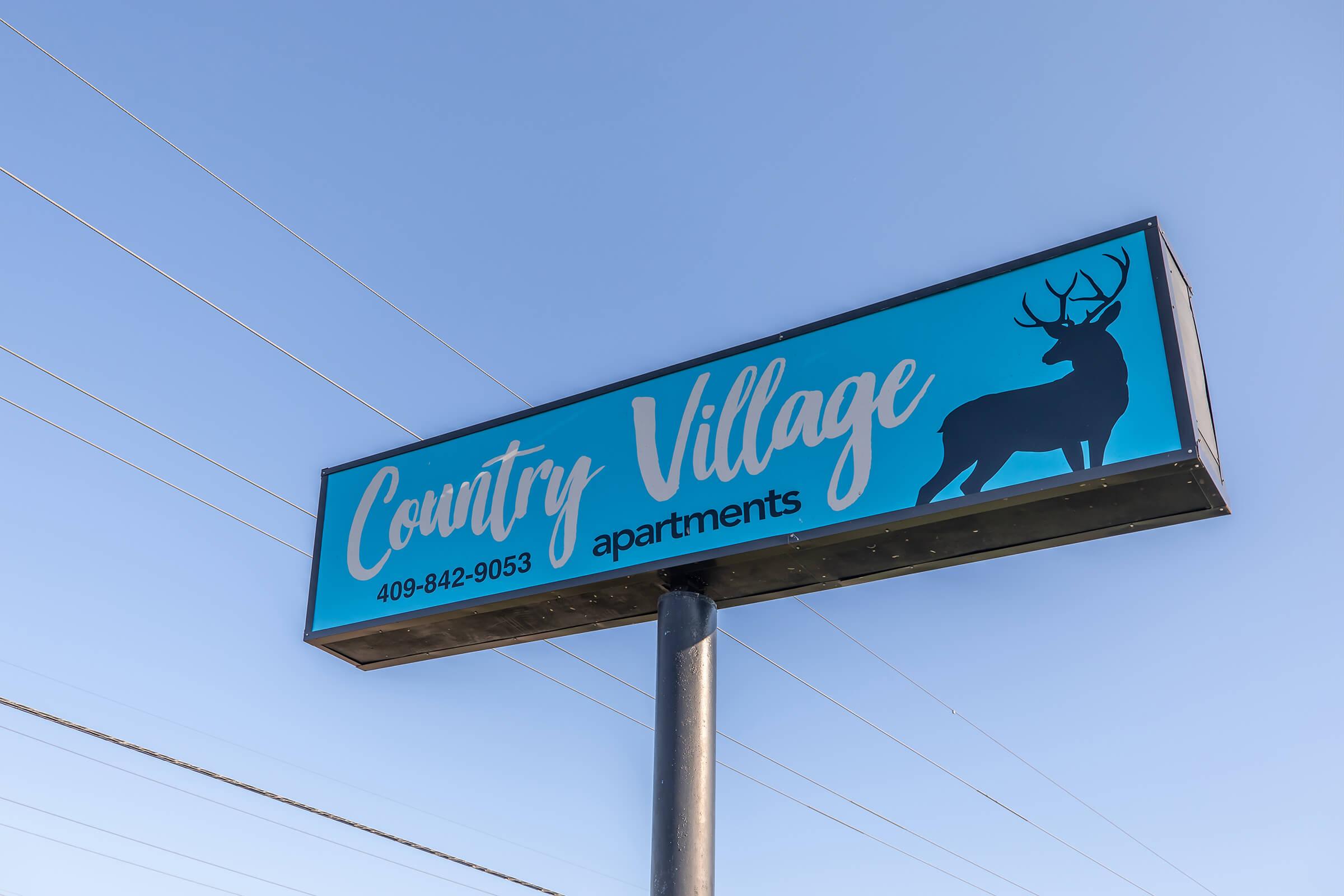 Sign for Country Village Apartments featuring a blue background, white text, and a silhouette of a deer. The sign includes a phone number for inquiries. The sky is clear with visible power lines in the background.