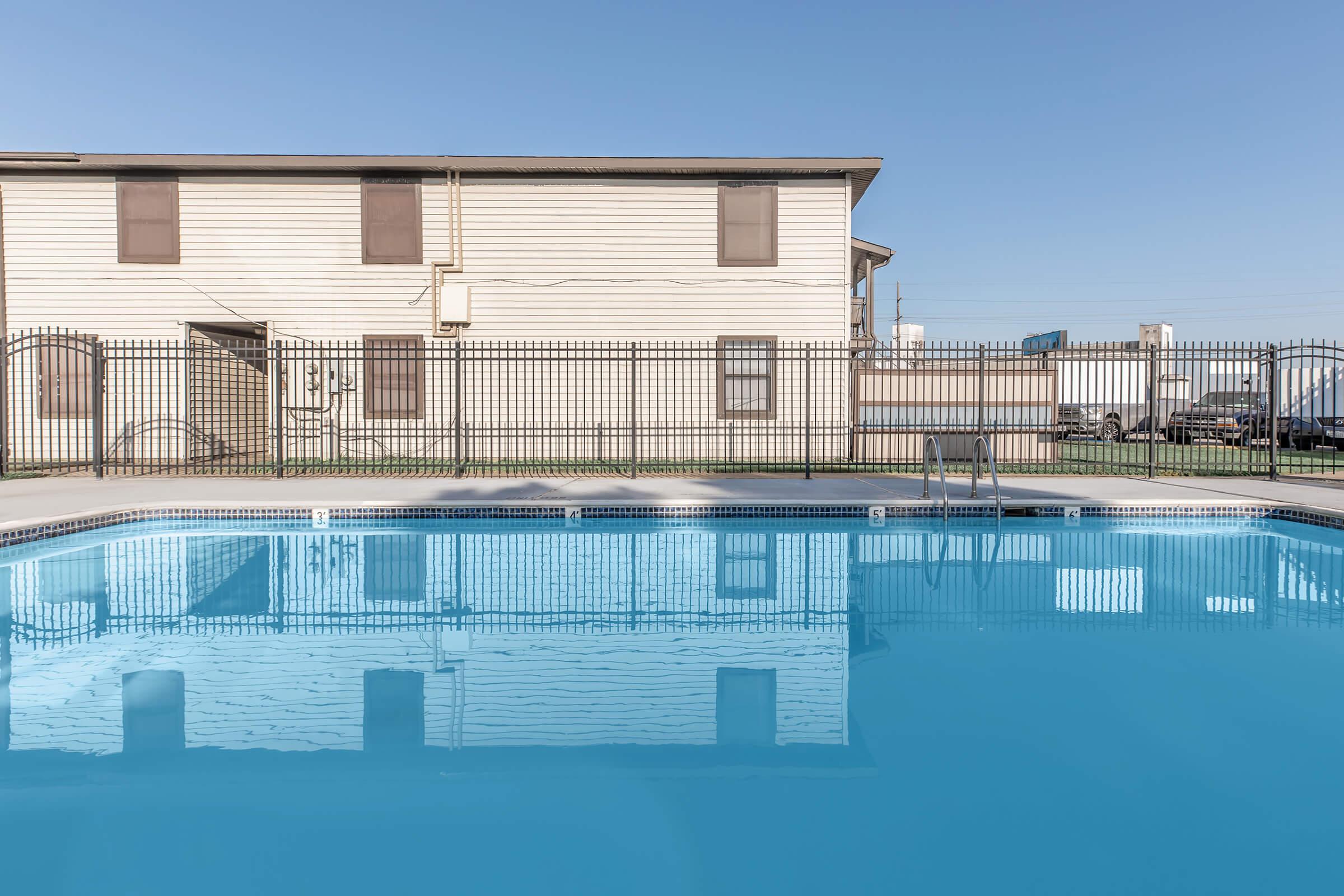 A clear blue swimming pool surrounded by a metal fence, reflecting a beige two-story building with closed windows in the background. The sky is bright and blue, and there are parked cars visible in the distance beyond the pool area.