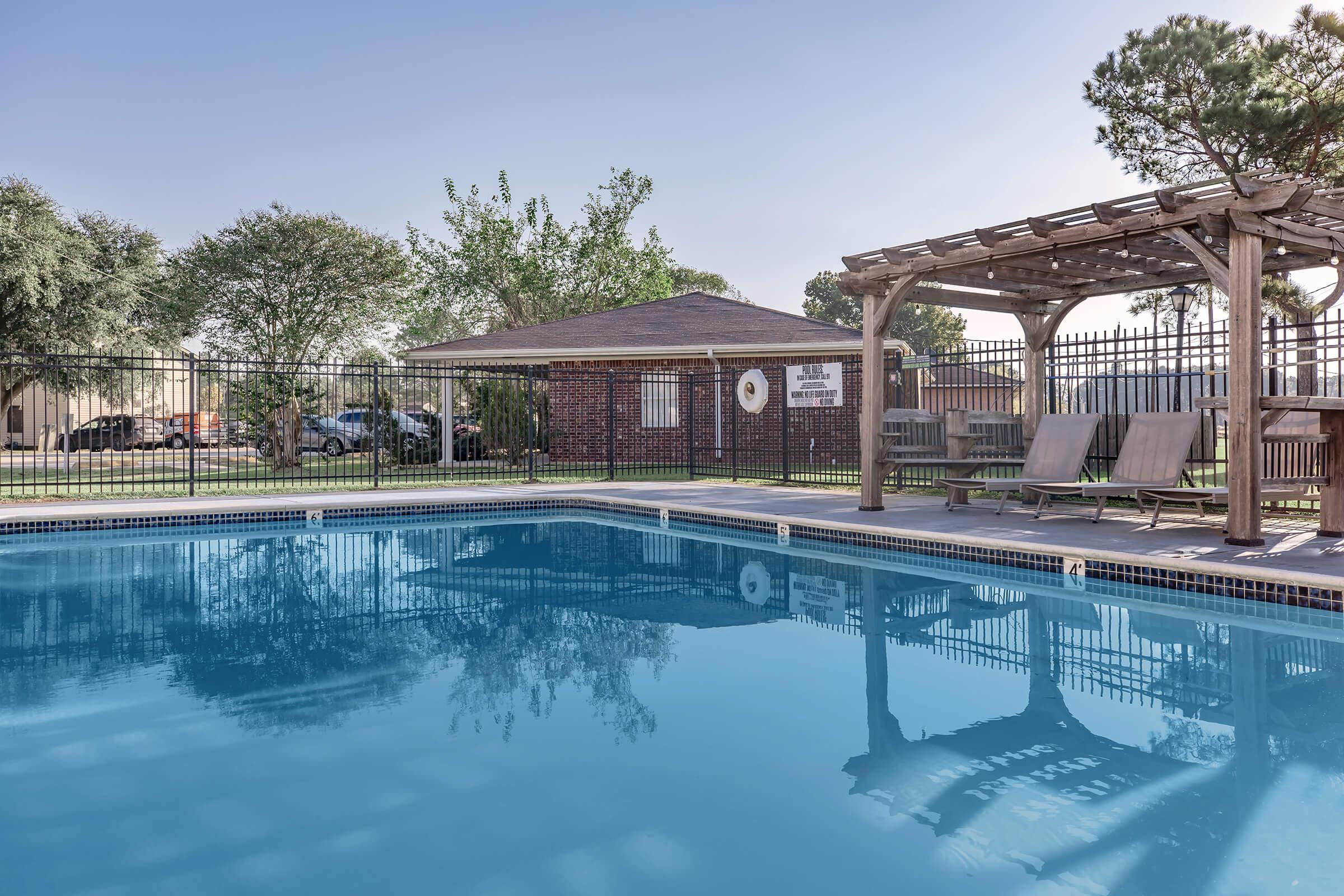 A swimming pool surrounded by a wooden deck with lounge chairs and a pergola. In the background, a well-maintained lawn and trees are visible. A building can be seen to the left, along with parked cars. The clear blue water reflects the sunny sky.