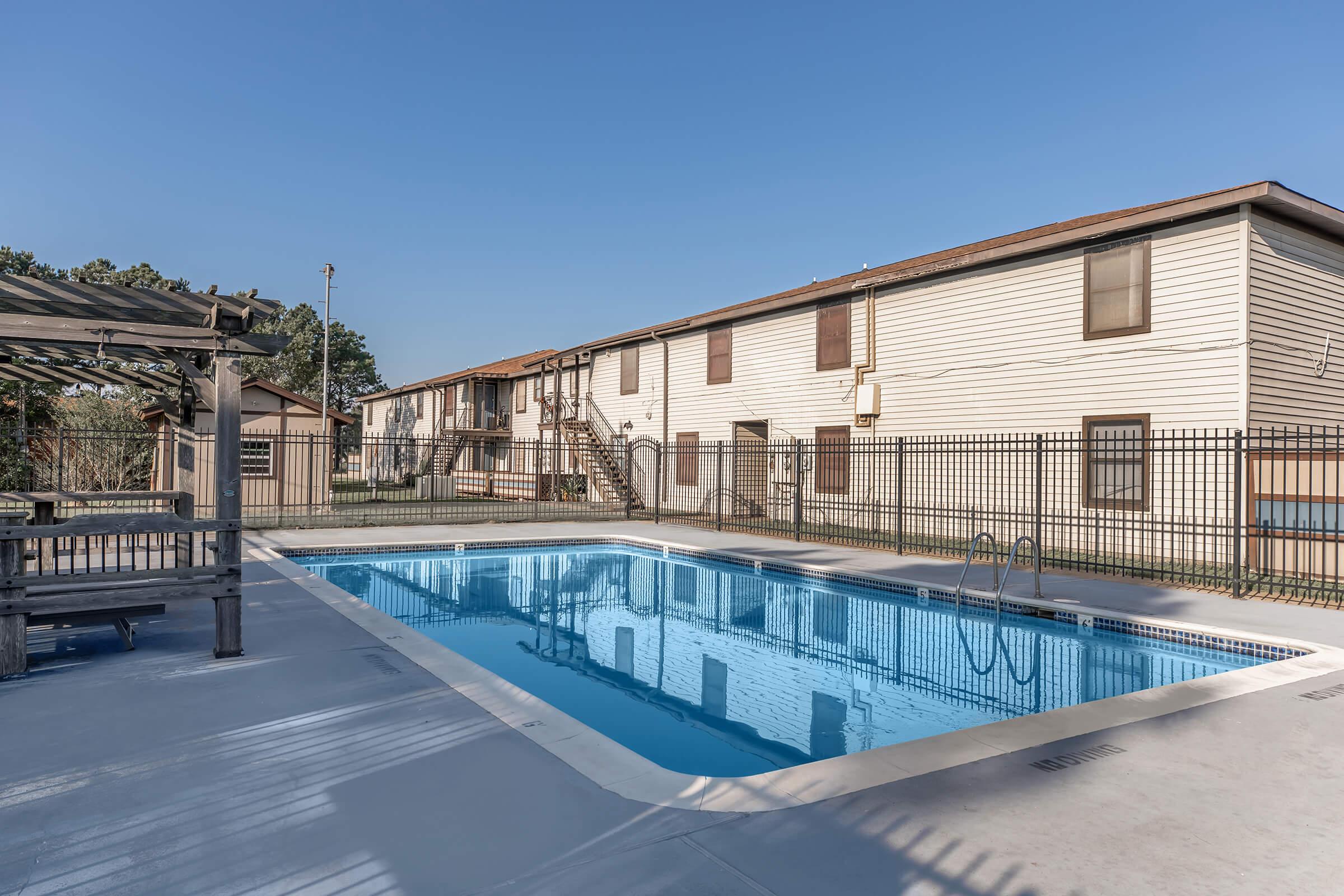 A swimming pool surrounded by a fence, with a building in the background. The pool area features a concrete deck and stairs leading into the water. The pool is clear and reflects the blue sky, while the building has several windows and a staircase visible. The scene is well-lit and inviting.