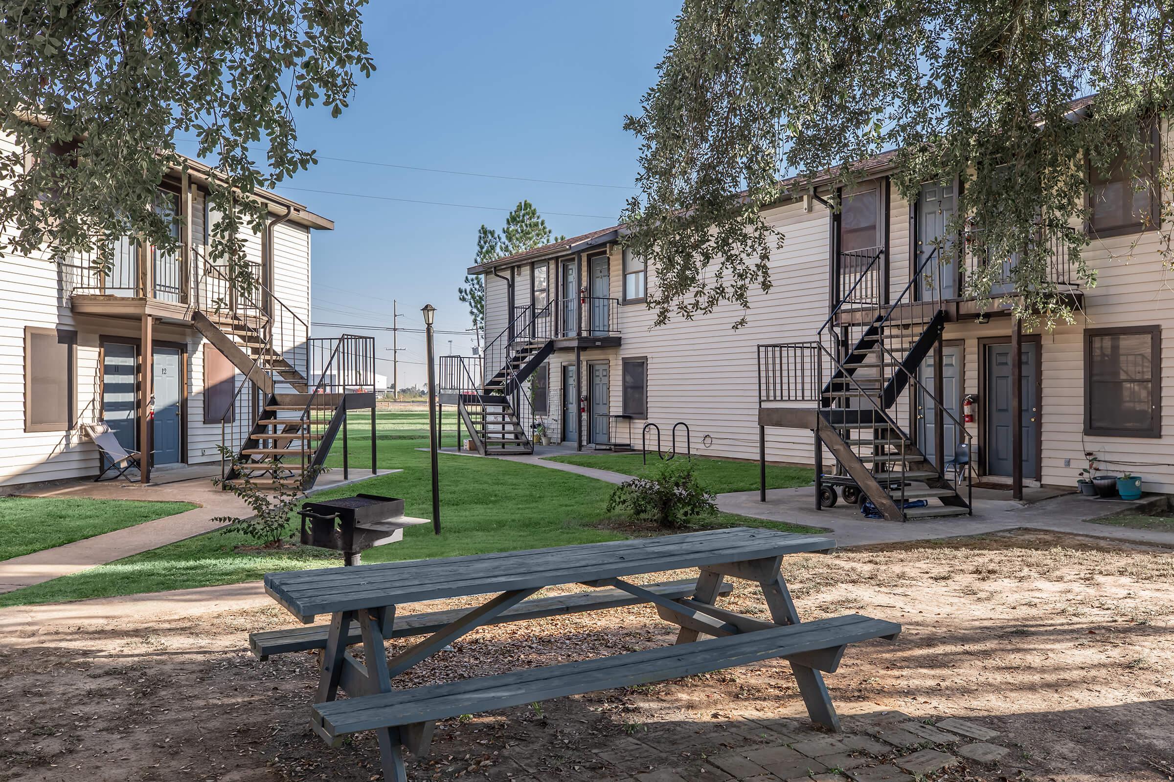 A view of a residential area featuring two sets of apartment buildings with outdoor staircases. In the foreground, there is a picnic table on a grassy area. The background shows clear blue skies and a few trees, contributing to a serene environment in this multi-family housing complex.