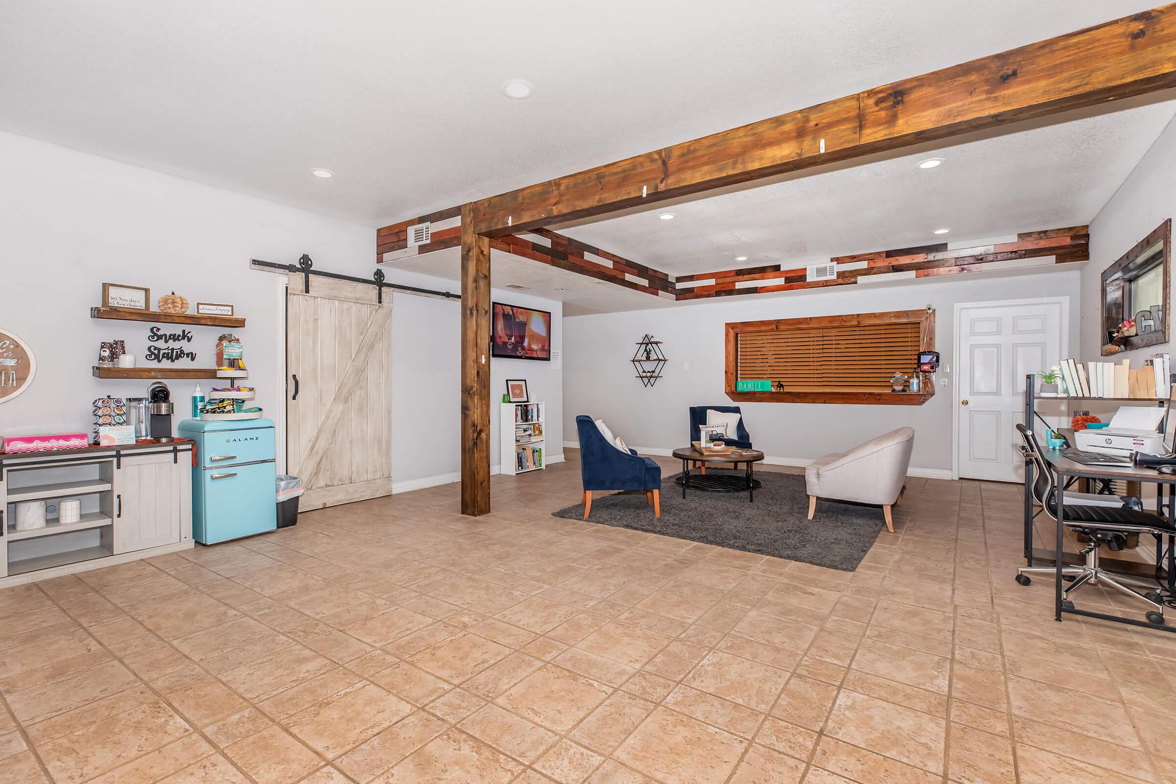 A spacious, modern living area featuring a sliding barn door, decorative wooden beams, and a tiled floor. There are two armchairs, a gray sofa, a small coffee table, and a retro-style blue refrigerator. A wall shelf and a desk with a chair provide additional storage and workspace.
