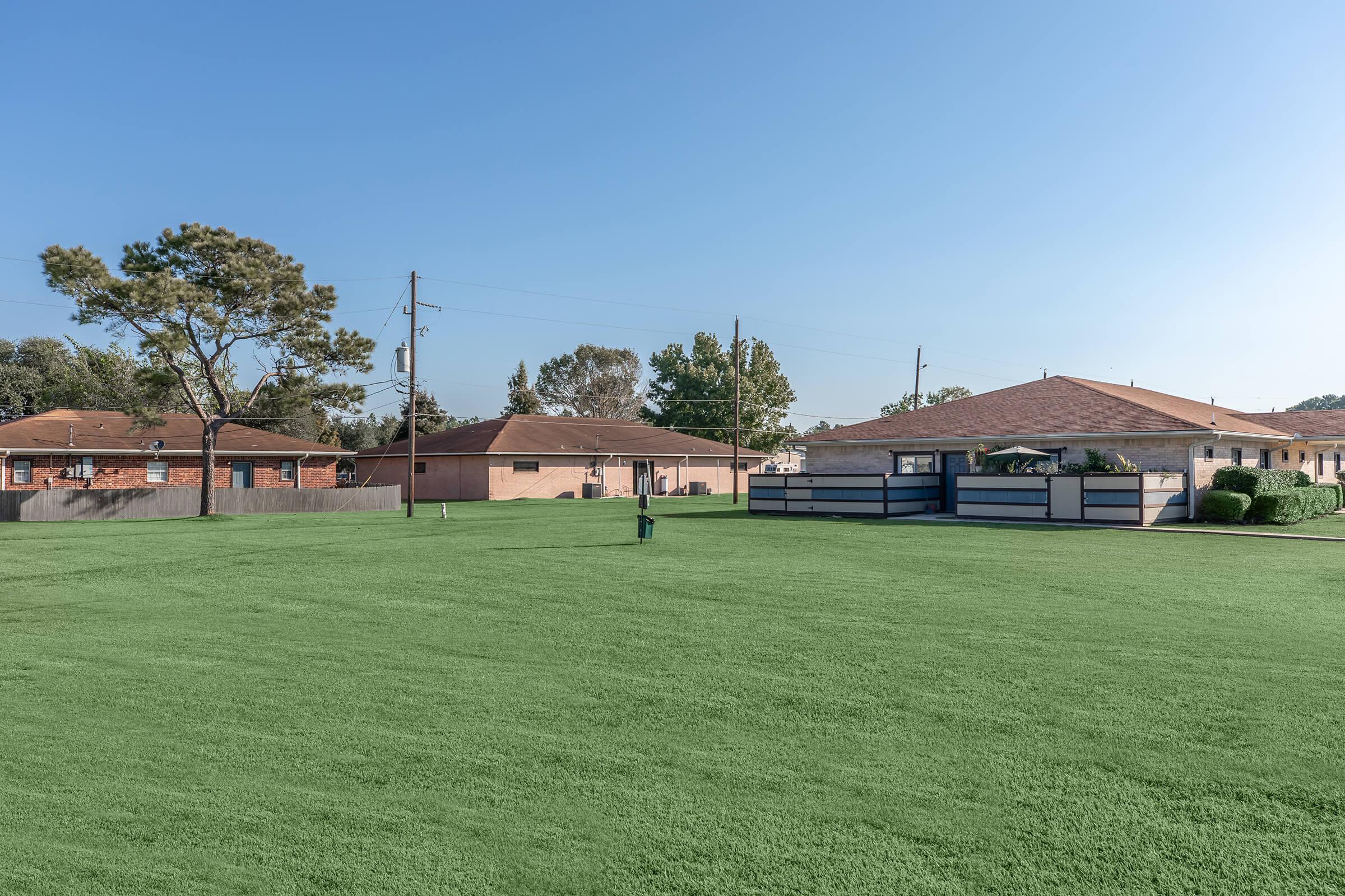 A sunny outdoor scene featuring a spacious green lawn bordered by residential buildings. The sky is clear and blue, showing a few trees in the background. Utility poles and wires can be seen, adding to the suburban setting. The area appears well-maintained and inviting.