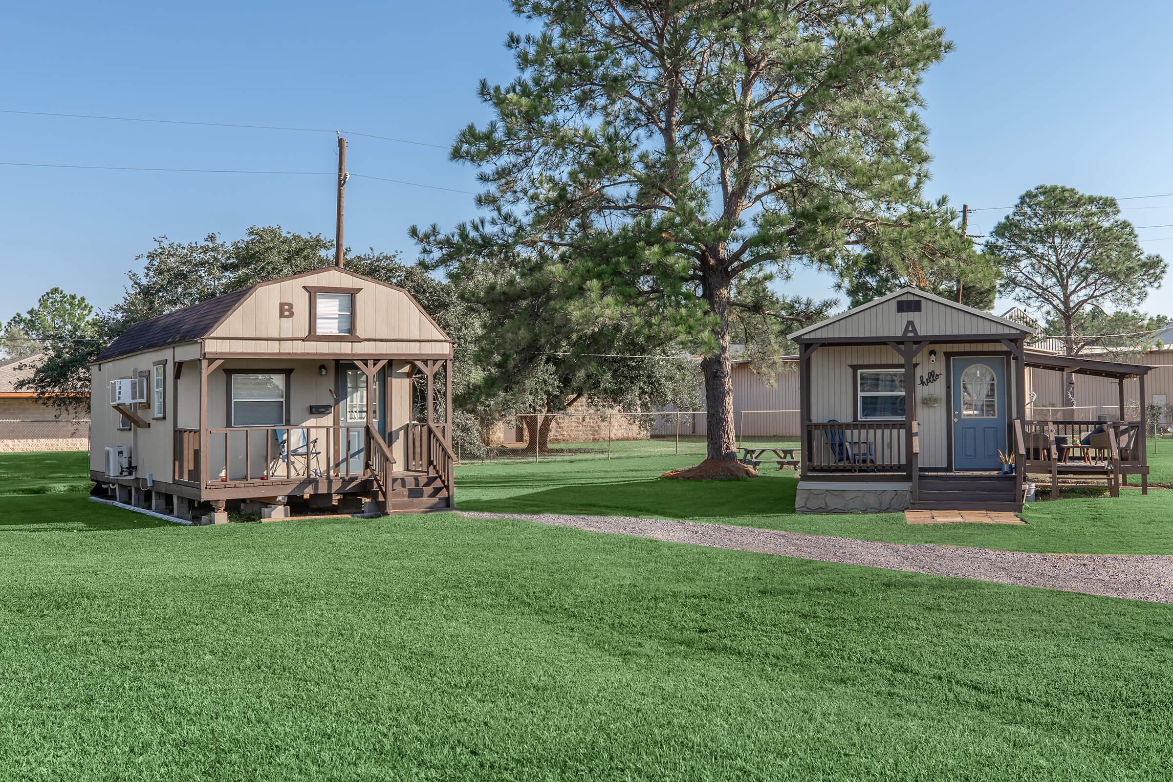 Two small cabins situated in a green outdoor area with a clear blue sky. The cabin on the left has a light brown façade and a deck leading up to the entrance. The cabin on the right features a darker exterior with a porch and seating area. Both cabins are surrounded by trees and a well-maintained lawn.