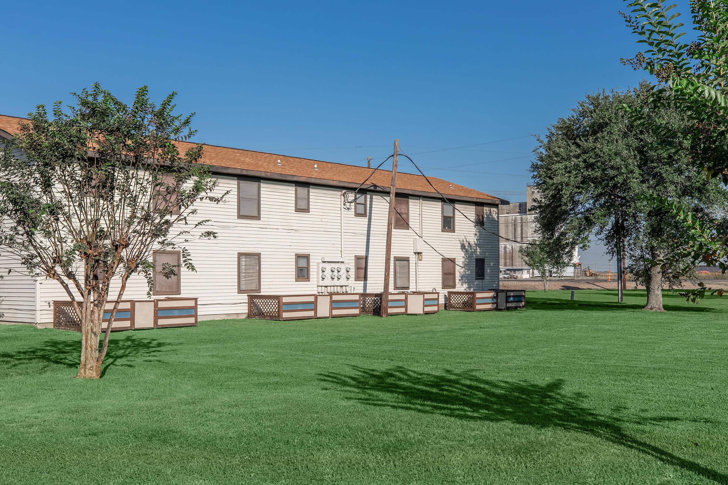 A two-story residential building with white siding and brown trim, surrounded by well-maintained green grass. There are small trees and bushes in the foreground, and a power pole in the background. The sky is clear and blue, indicating a sunny day.