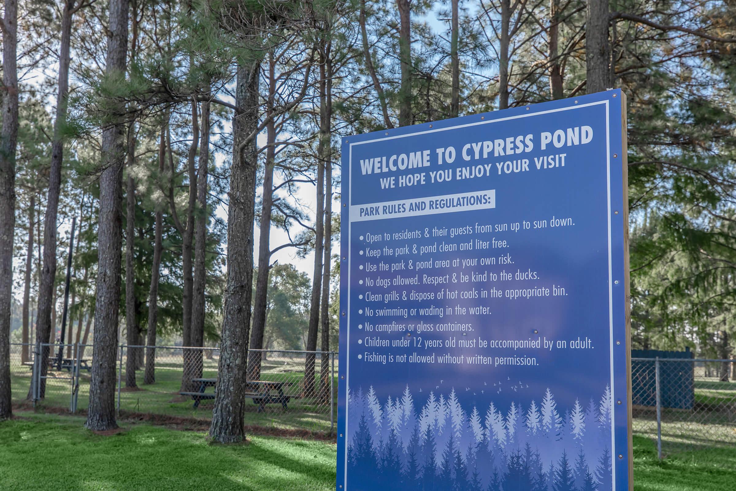 A sign welcoming visitors to Cypress Pond, outlining park rules and regulations. It includes guidelines such as keeping the park clean, respecting wildlife, and restrictions on swimming and fishing. The background features a wooded area with trees and picnic tables.