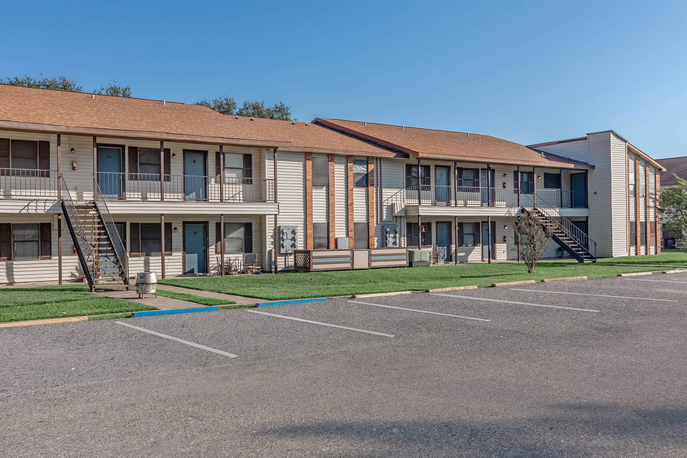 A row of two-story apartment buildings with beige siding and brown roofs, featuring staircases leading to the upper units. The foreground shows a parking lot with marked spaces and green grass, under a clear blue sky.