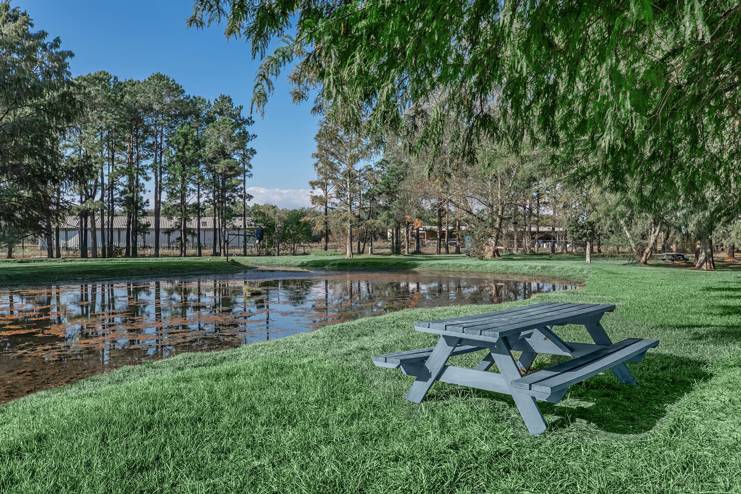 A serene landscape featuring a gray picnic table near a calm pond, surrounded by lush green grass and tall trees. The sky is clear with a few clouds, and the reflection of the trees is visible on the water's surface, creating a peaceful outdoor setting.