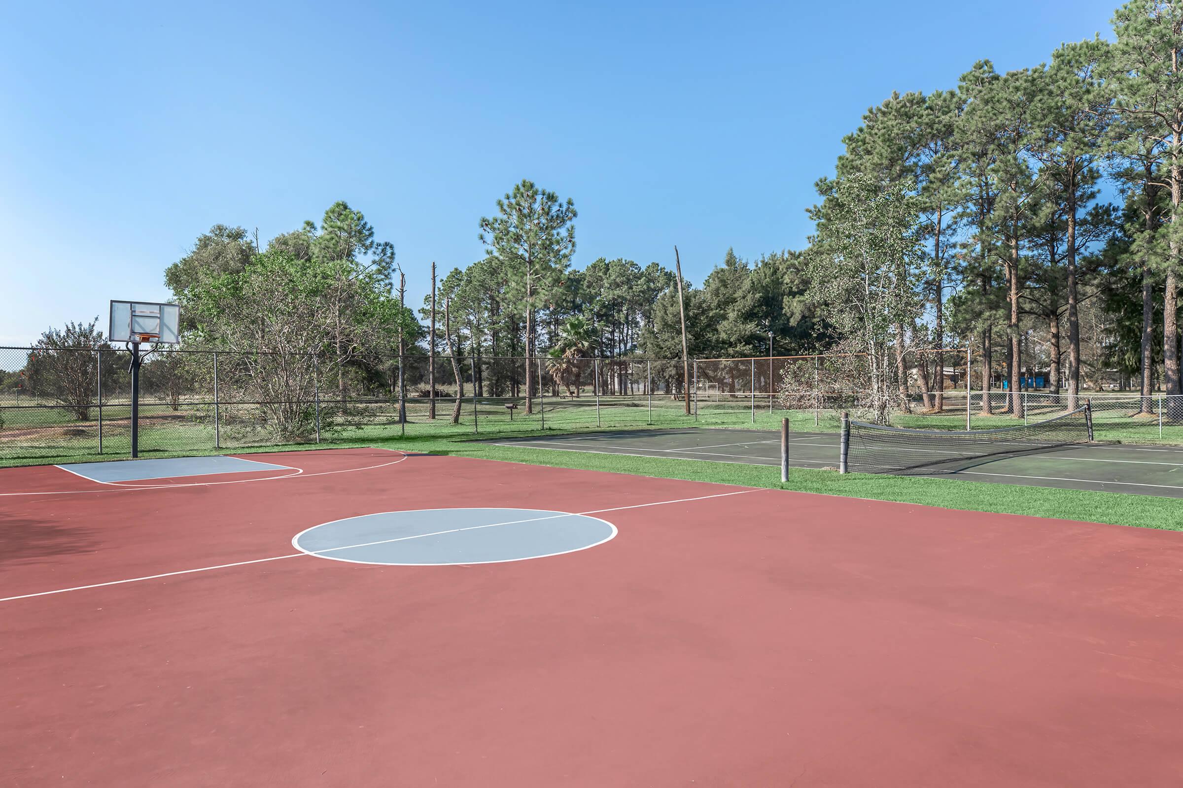 A view of a basketball court with red and gray markings, set against a backdrop of green trees and a blue sky. A second tennis court is visible in the background, featuring a grassy area and fencing, providing a serene outdoor recreational space.