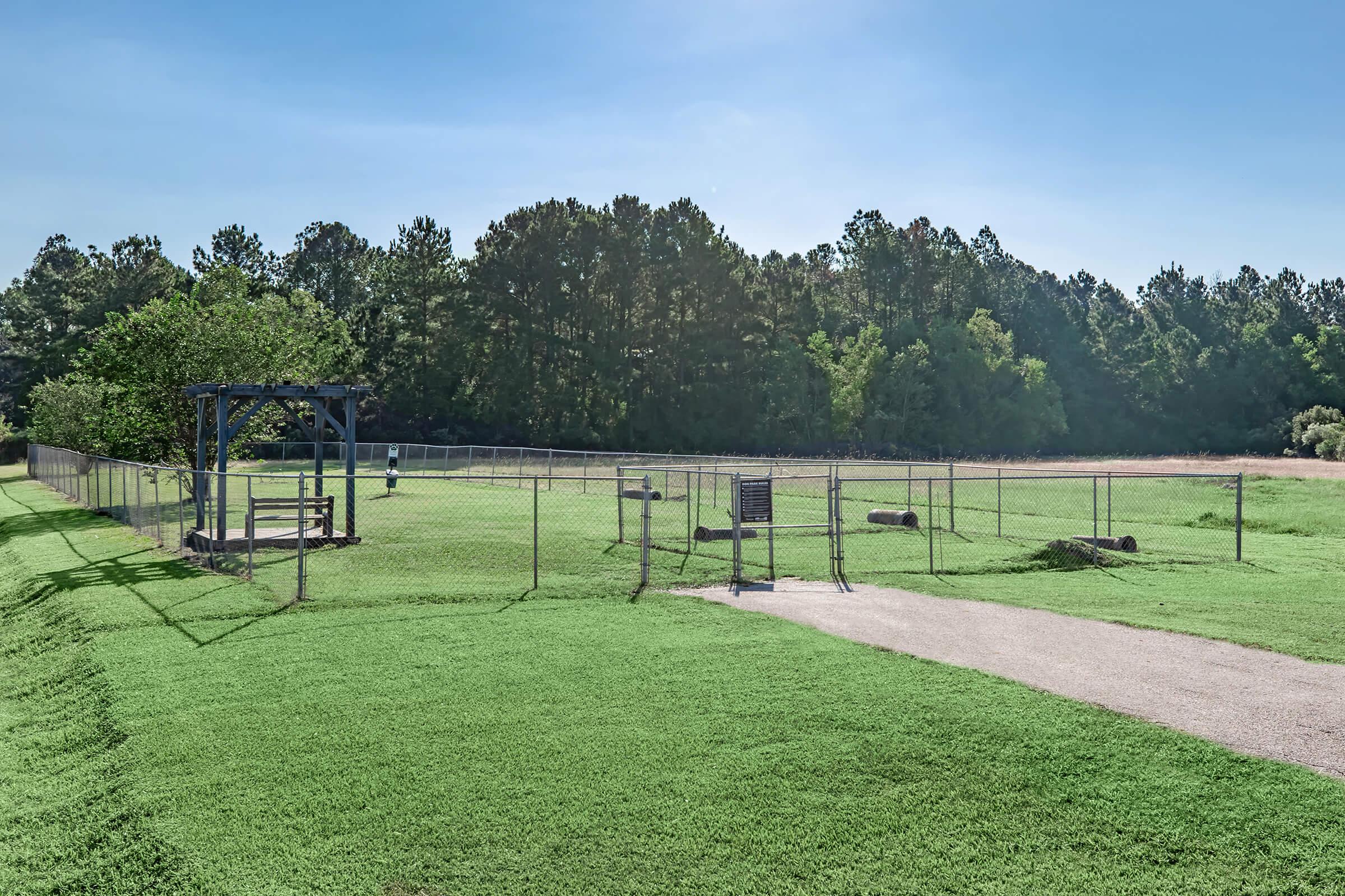 A fenced open area with a grassy field and a gravel path. There are trees in the background and a few wooden structures within the enclosure, possibly for seating. The sky is clear, suggesting a sunny day. A sign is visible near the entrance of the fenced area.