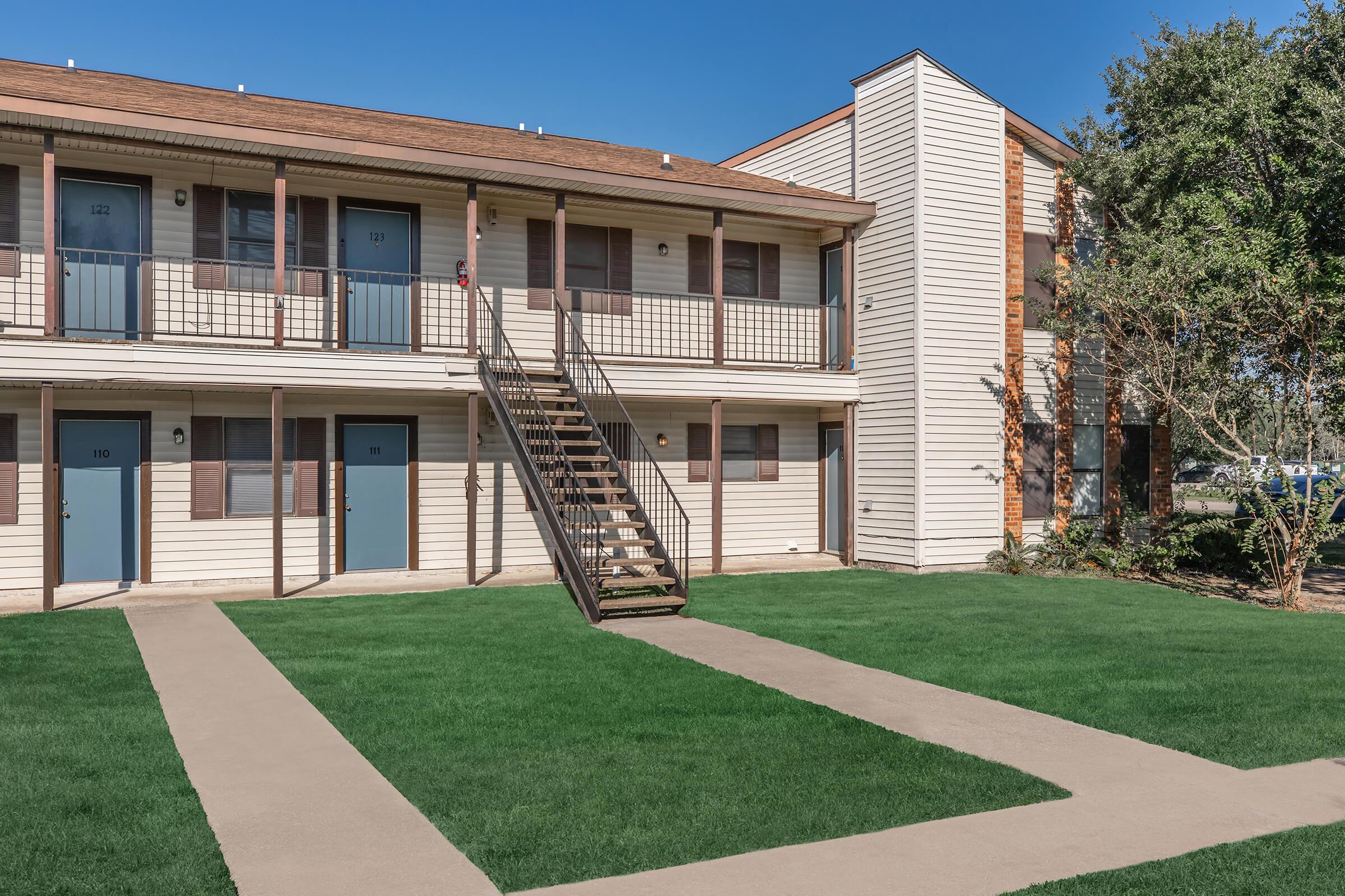 A two-story apartment building with multiple units, featuring a staircase leading to the second floor. The exterior is light-colored with wooden accents, and the ground is covered in green grass. Pathways lead to the entrances of the apartments, and the sky is clear and blue.