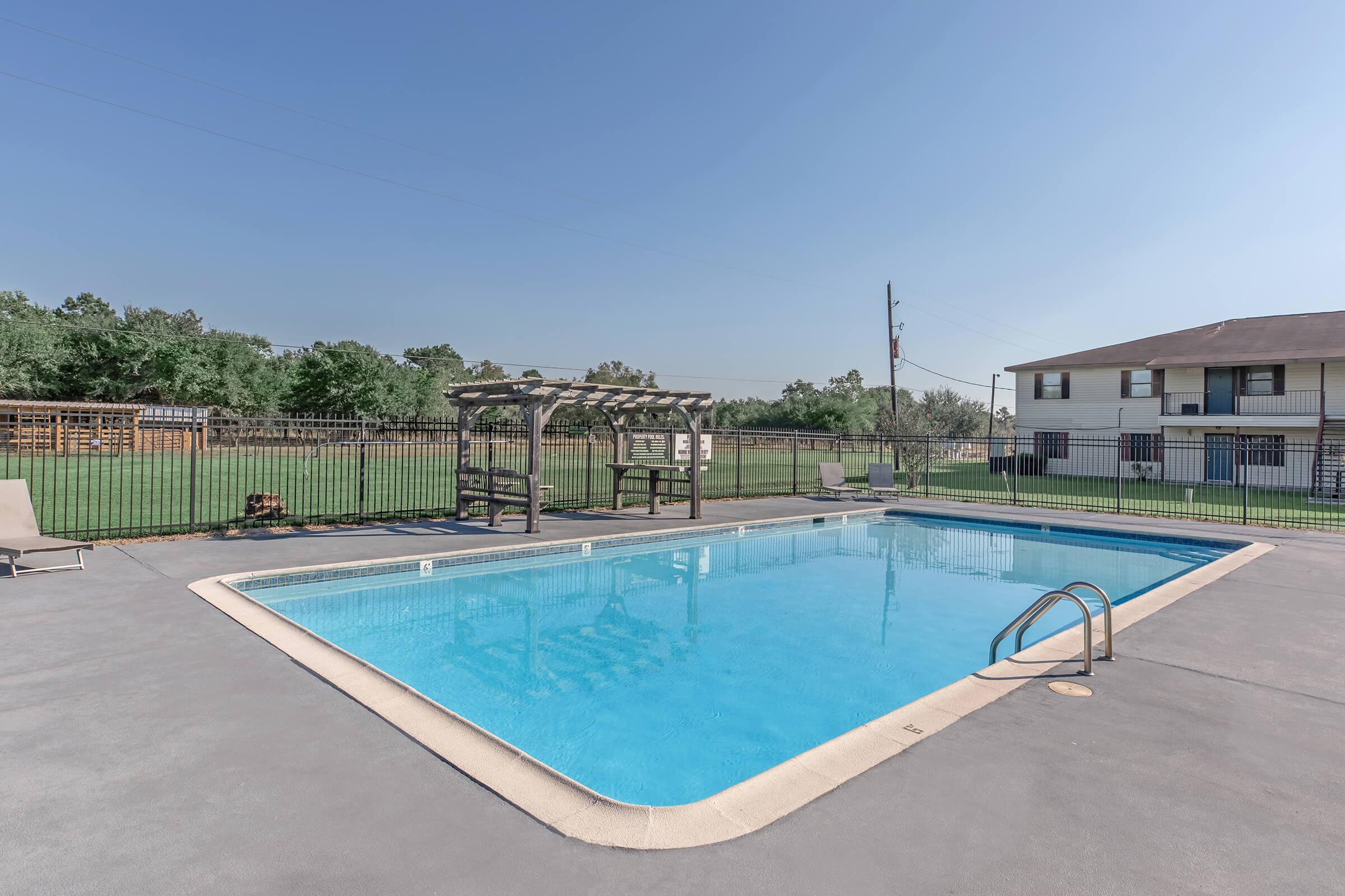 A clear blue swimming pool surrounded by a concrete deck. There are lounge chairs on the sides, a pergola near the pool area, and a grassy field in the background. A two-story building is visible at the rear, with utility poles in the distance and a clear blue sky overhead.