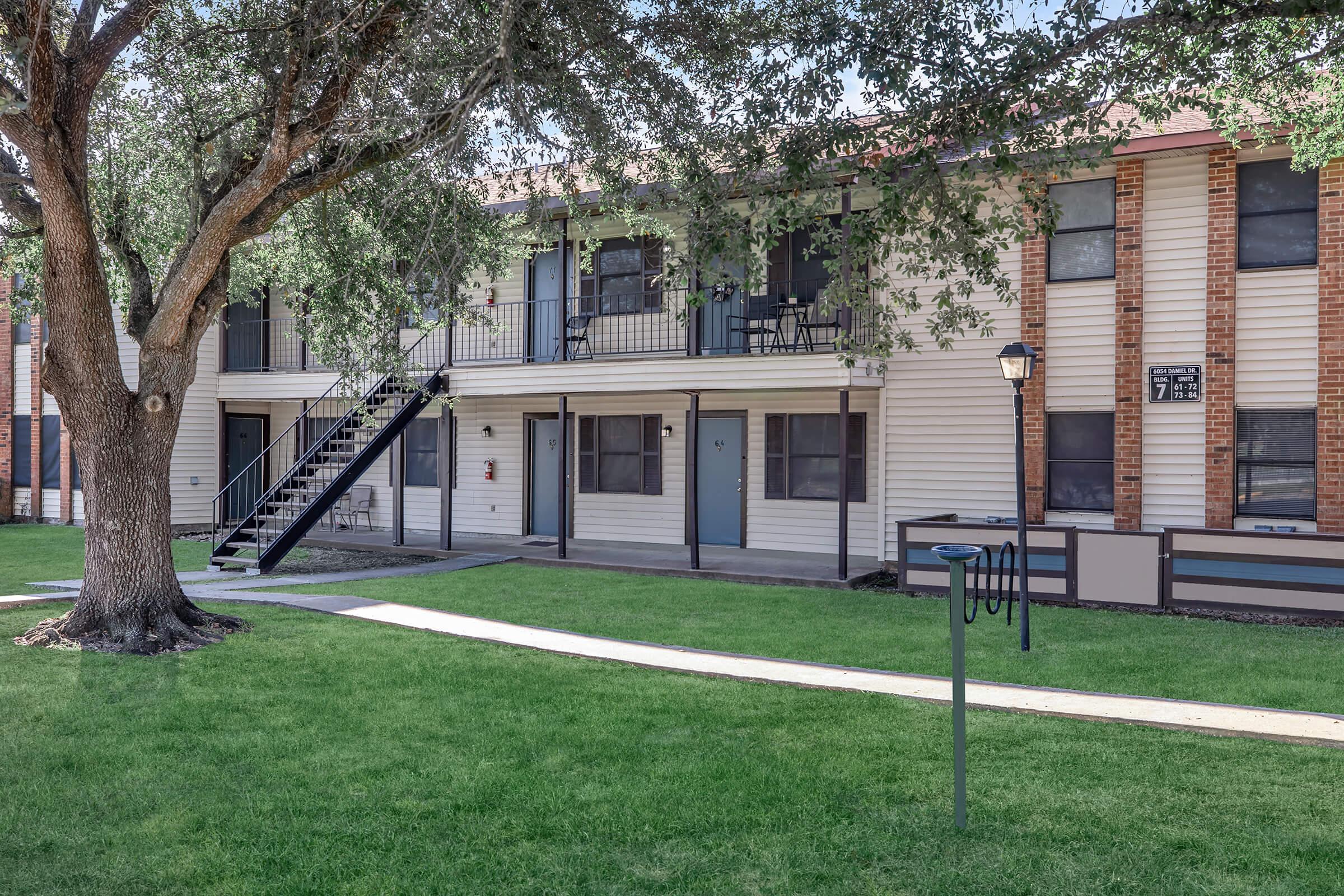 A view of a two-story apartment building with a central staircase. The building features multiple front doors, large windows, and is surrounded by green grass and a tree. The setting appears well-maintained, with a clear blue sky above.