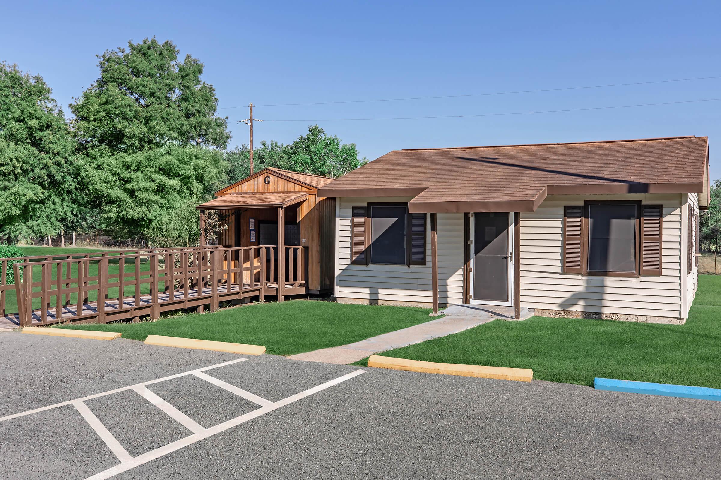 A small one-story building with a sloped roof, featuring brown trim and large windows, sits alongside a wooden ramp leading to a smaller structure. The area is well-maintained with green grass and trees in the background. A parking lot is visible in the foreground with marked spaces.