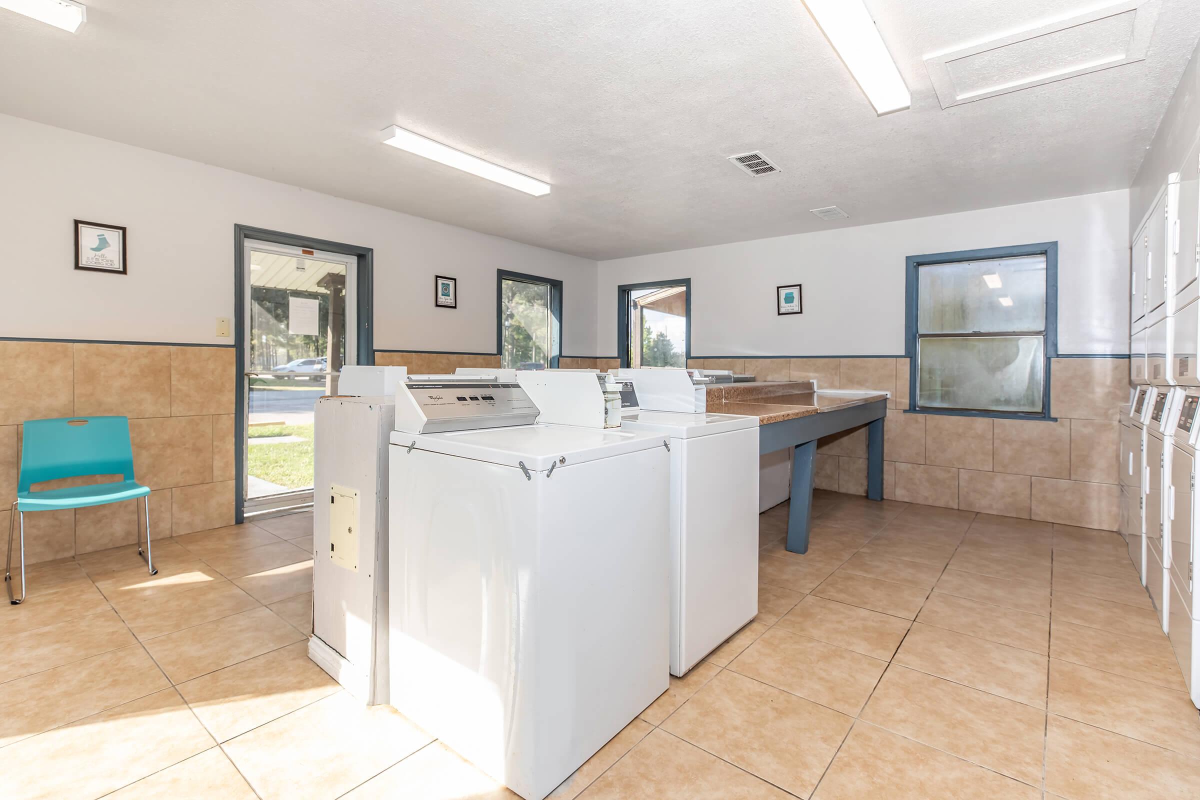 A clean and well-lit laundry room featuring several washing machines, a folding table, and lockers. The space has tiled flooring, beige walls, and windows that allow natural light. A blue chair is positioned against the wall, and there are framed pictures on the walls.