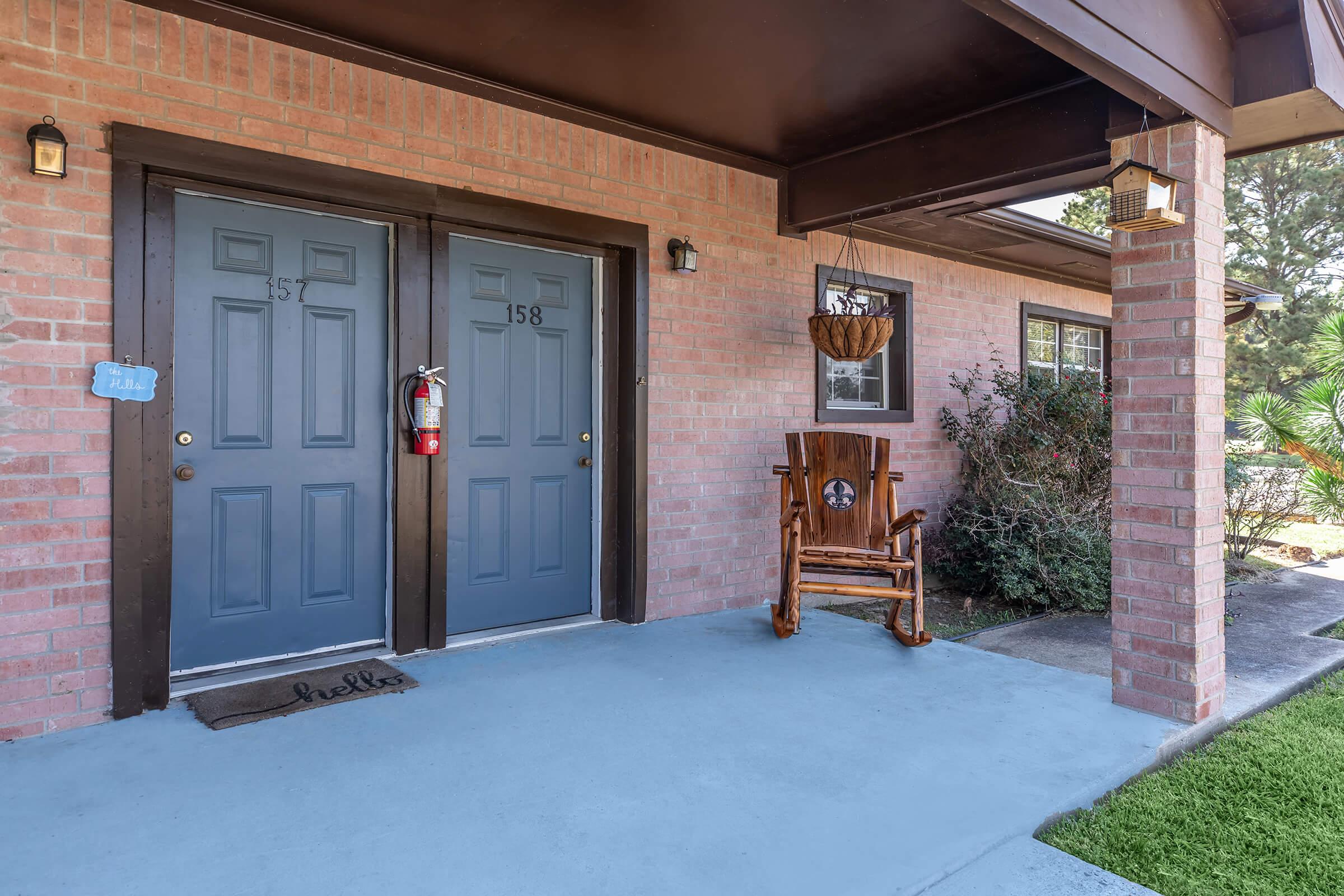 Entrance of a residential building featuring two blue doors labeled "157" and "158." There is a wooden rocking chair on the porch, a fire extinguisher mounted on the wall, and a doormat that says "Hello." Surrounding the entrance are shrubs and palm trees, creating a welcoming atmosphere.