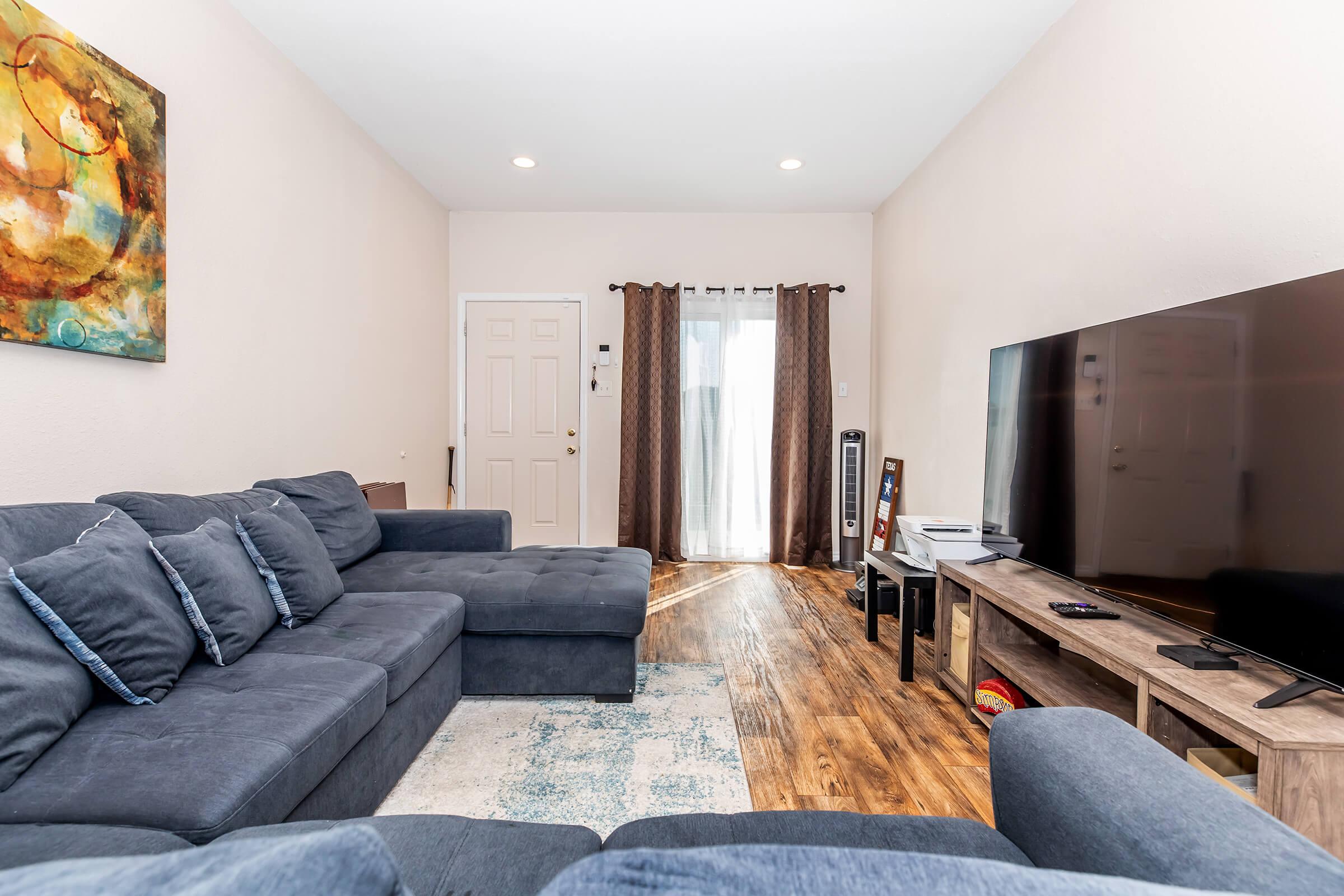 Living room with a large, gray sectional sofa, wooden TV stand, and a flat-screen TV. Natural light enters through the window covered with dark curtains. A door leads outside, and a light-colored area rug is placed on the wooden floor, creating a cozy atmosphere.