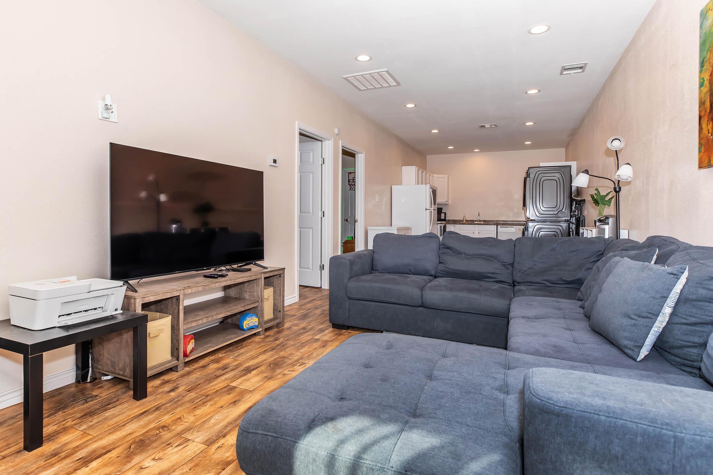 Living room featuring a large dark gray sectional sofa and a flat-screen TV on a wooden stand. In the background, there is a modern kitchen visible with white appliances. The walls are painted light beige, and the floor has wood-like laminate. A colorful painting hangs on the wall, adding a vibrant touch to the space.