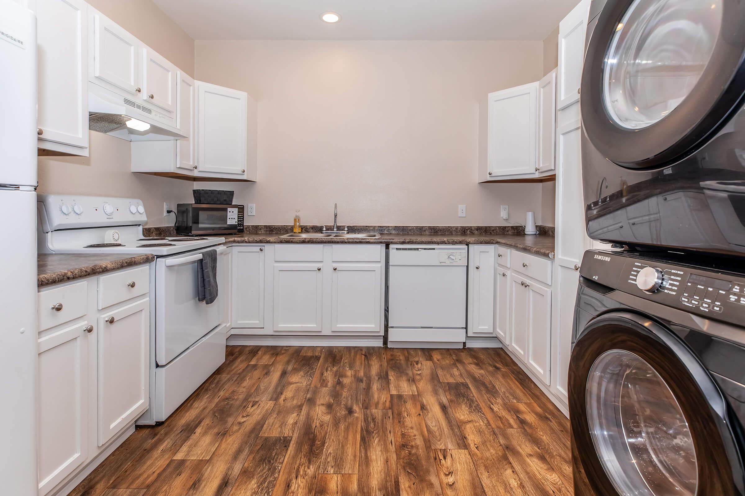 A modern kitchen featuring white cabinets, a brown countertop, and stainless steel appliances. The space includes a stove, microwave, and dishwasher, along with a washer and dryer stacked next to each other. The floor is made of hardwood laminate, creating a warm and inviting atmosphere.