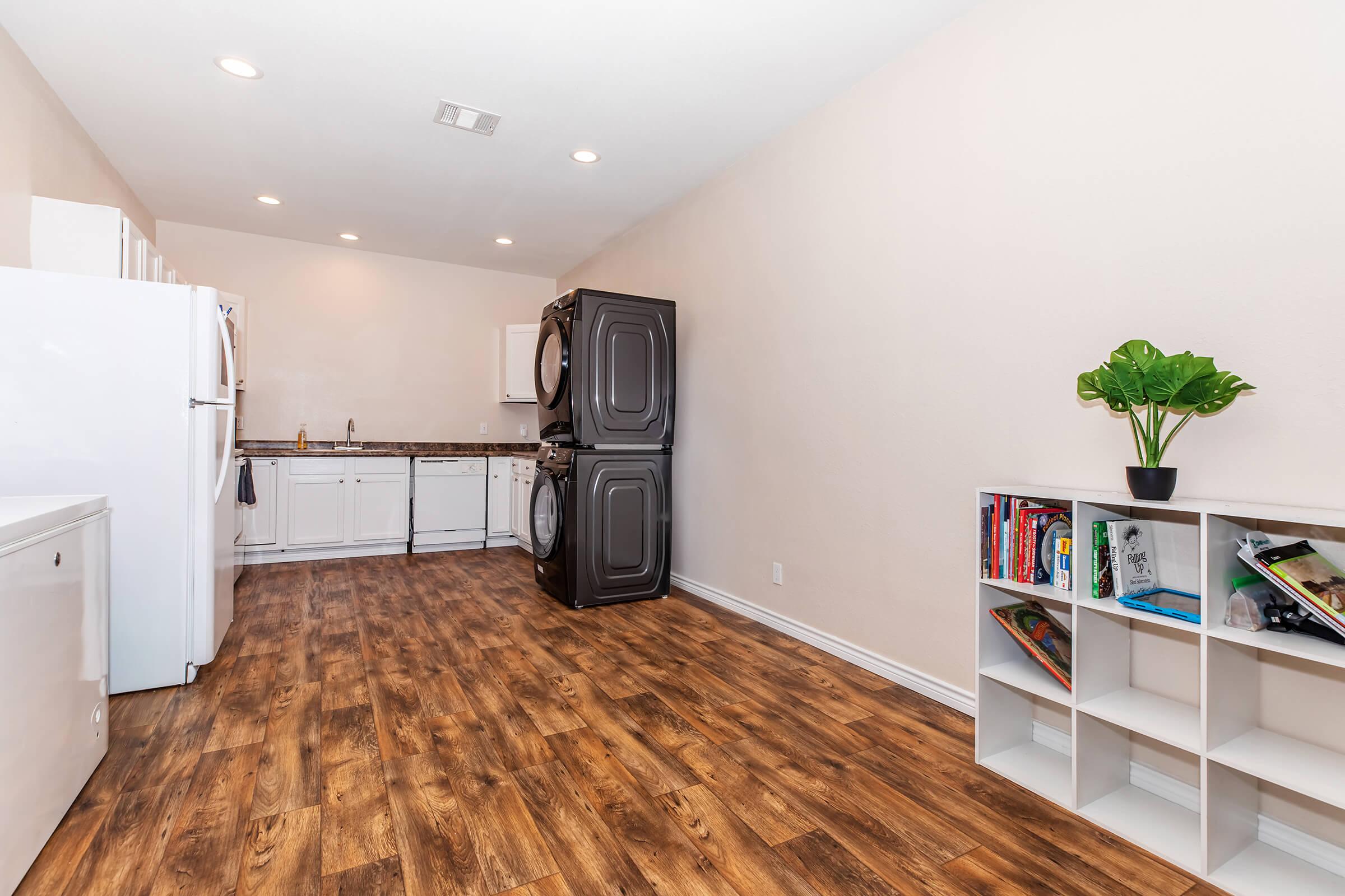 A spacious laundry area featuring a washer and dryer stack, with a modern kitchen in the background. The kitchen has white cabinets and a countertop, while the floor is finished in wood-like laminate. A small shelf holds books and a potted plant adds a touch of greenery to the room.