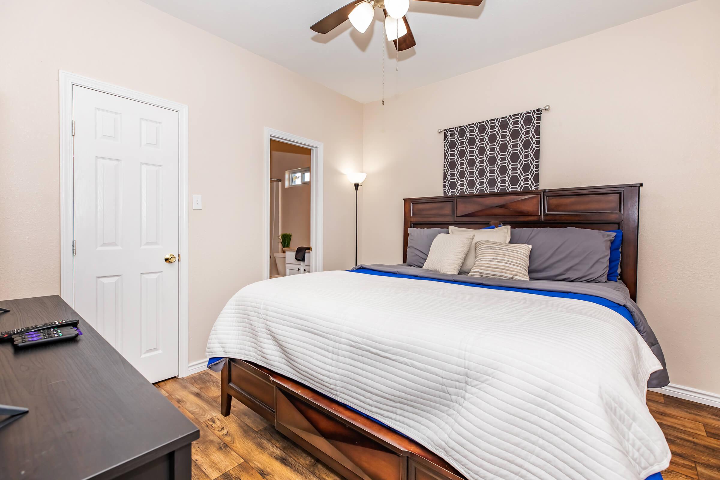 A cozy bedroom featuring a large bed with gray and blue bedding, a ceiling fan, a wooden headboard, and a dark wooden dresser. There's a large window with light curtains and a lamp in the corner, creating a warm atmosphere. A door leads to another room in the background.
