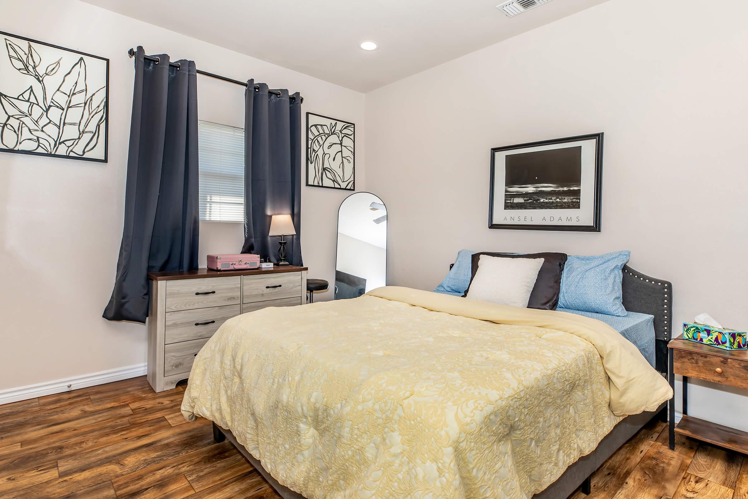 A cozy bedroom featuring a neatly made bed with a yellow blanket, decorative pillows, and a gray upholstered headboard. There are two black curtains framing a window, a dresser with a mirror, and framed botanical art on the walls. The room has warm wooden flooring and a soft, inviting atmosphere.