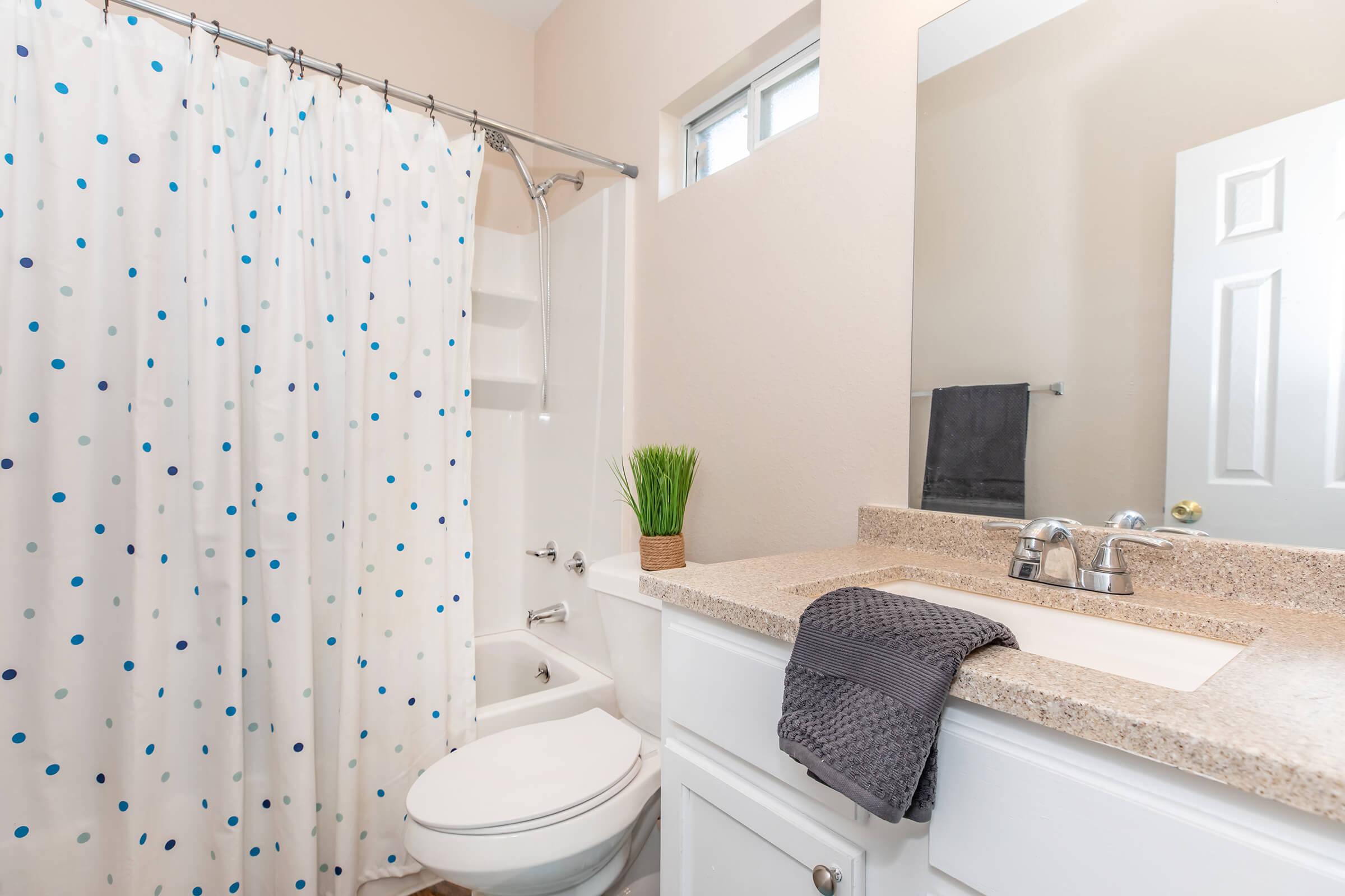 A well-lit bathroom featuring a modern design. There is a white shower curtain with blue dots, a white bathtub, and a vanity with a beige countertop. A small potted plant adds a touch of greenery, while a gray towel is neatly placed on the counter. A mirror hangs above the sink, enhancing the light space.
